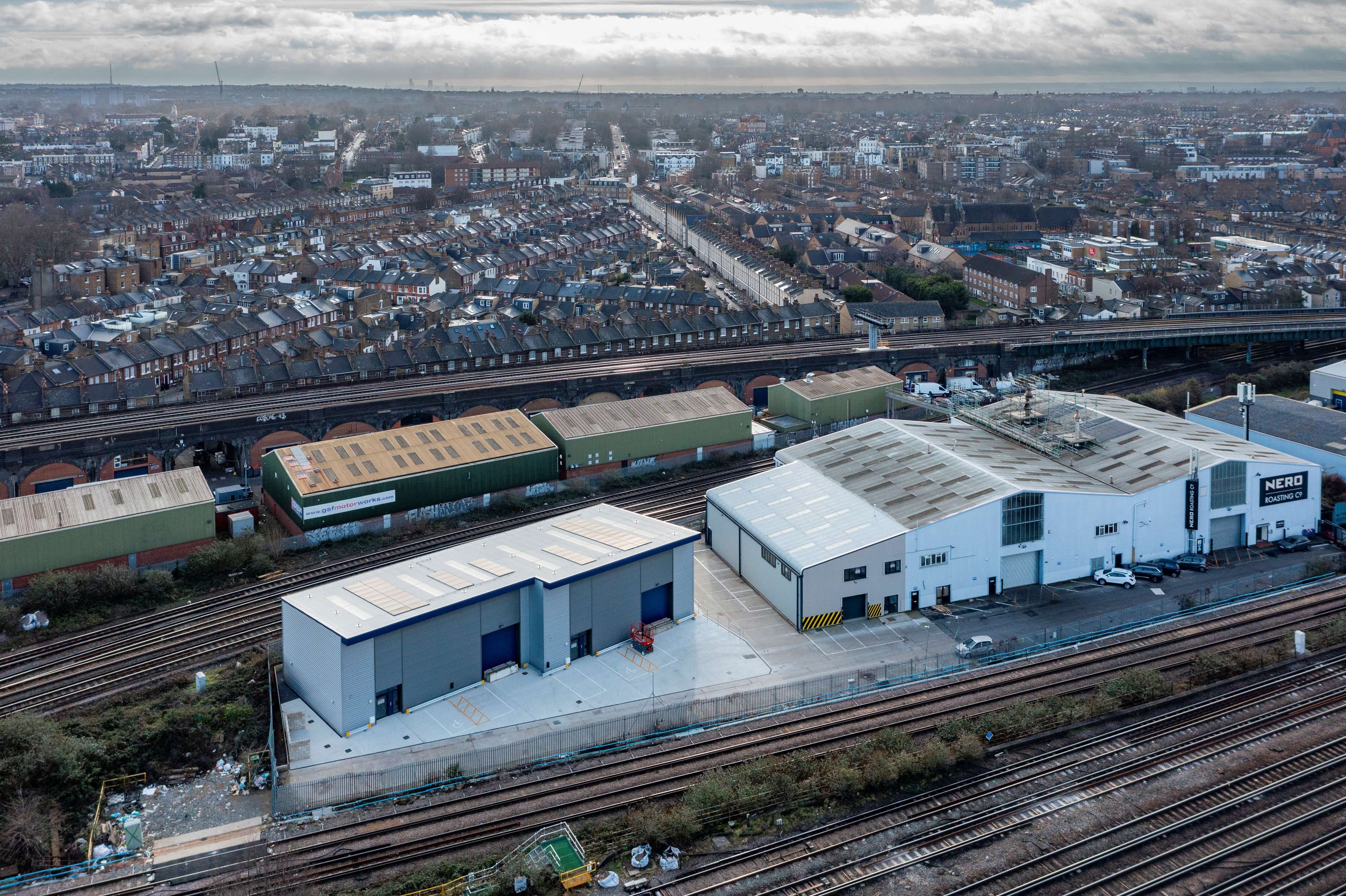 Aerial view of a warehouse complex beside railroad tracks in an urban area, with residential neighborhoods and city infrastructure in the background.
