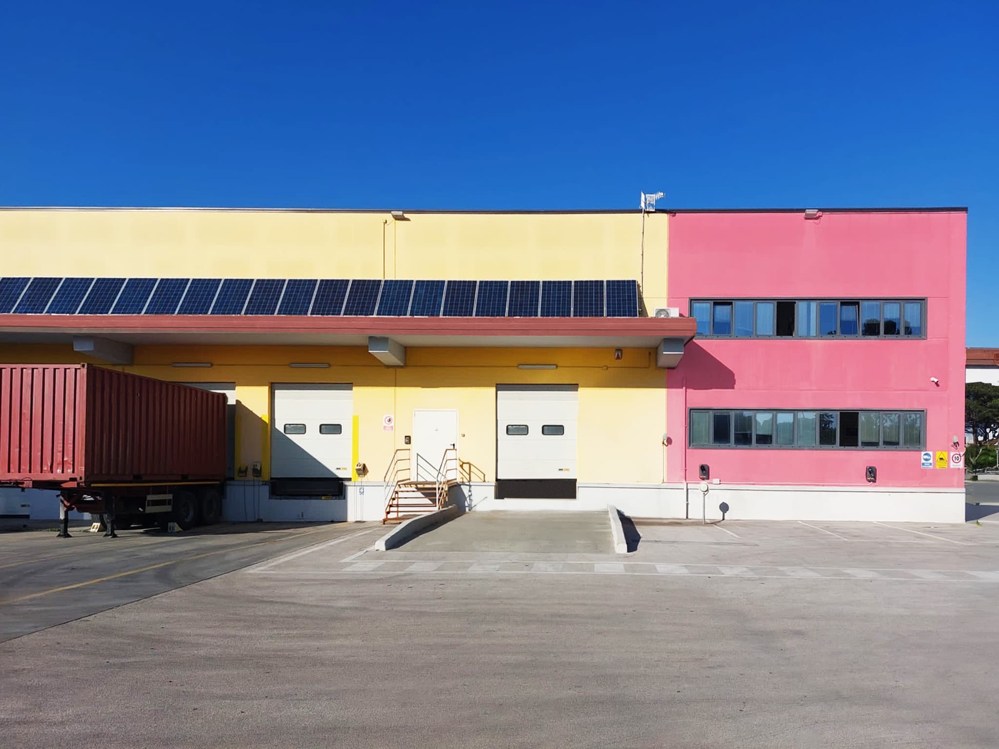 A yellow and pink industrial building with solar panels on the roof and a parked truck. There are two large garage doors and clear blue skies in the background.