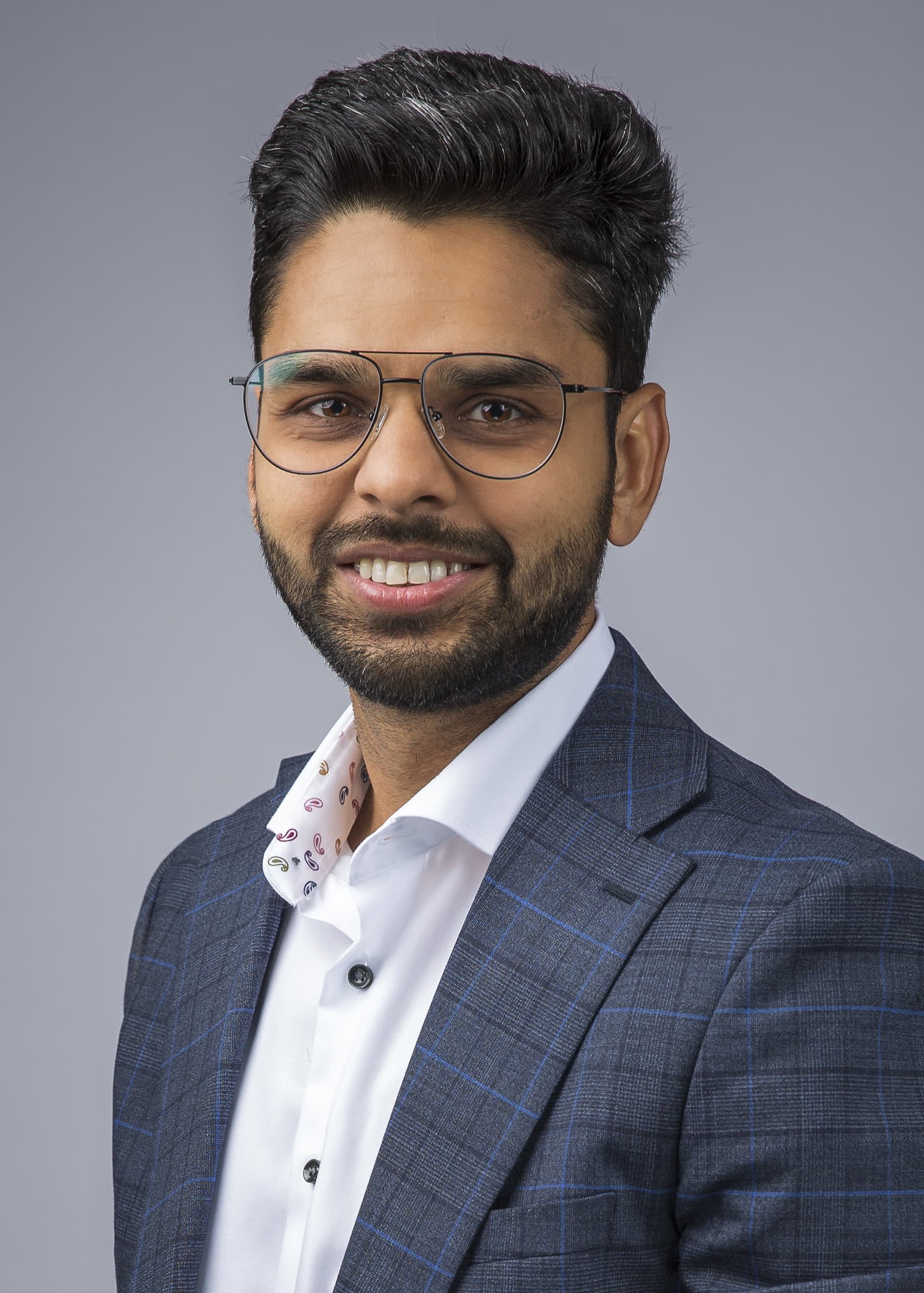 Person wearing glasses, a white shirt, and a blue checkered suit jacket, smiling against a plain gray background.