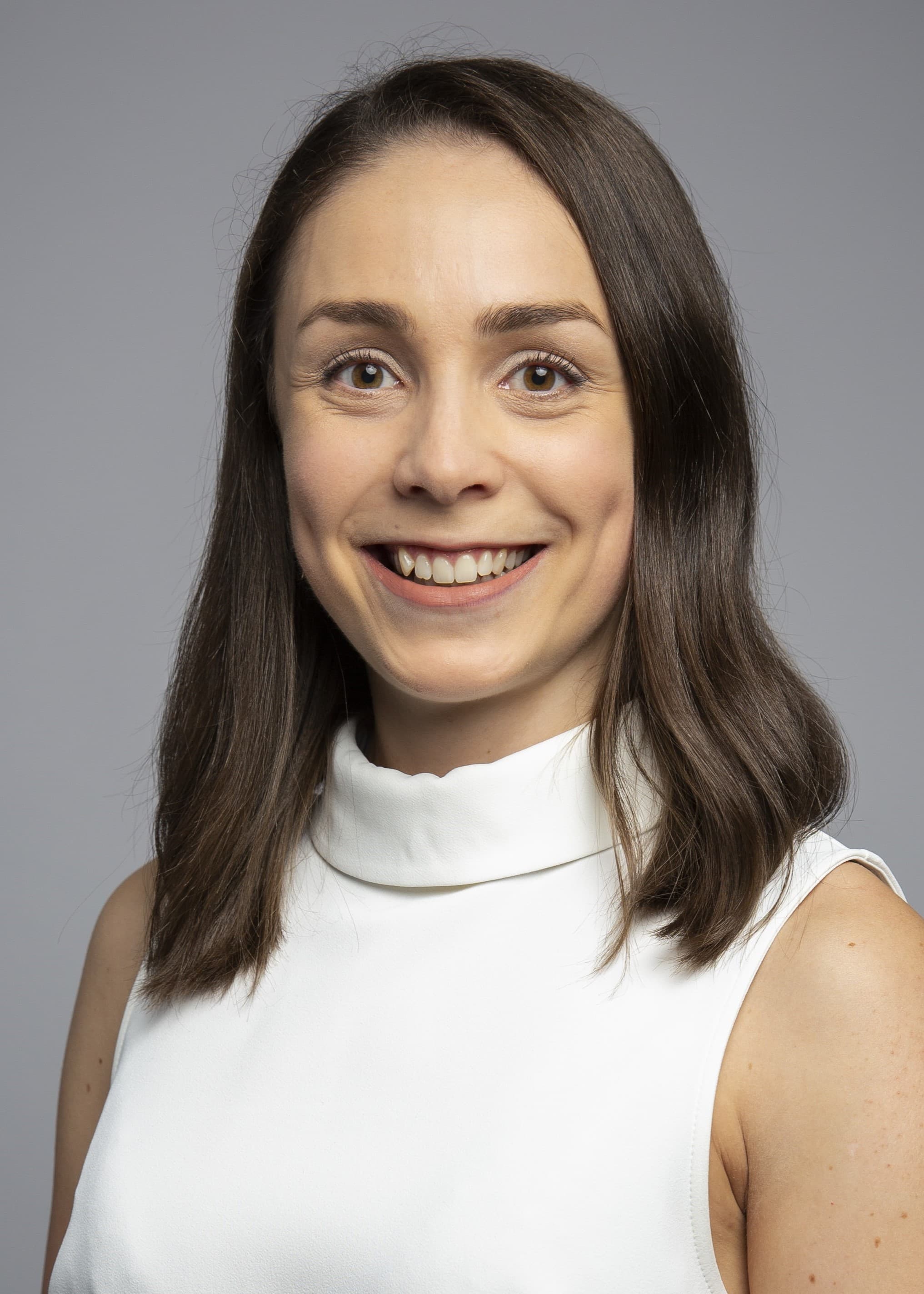 A person wearing a sleeveless white top smiles at the camera against a plain gray background.