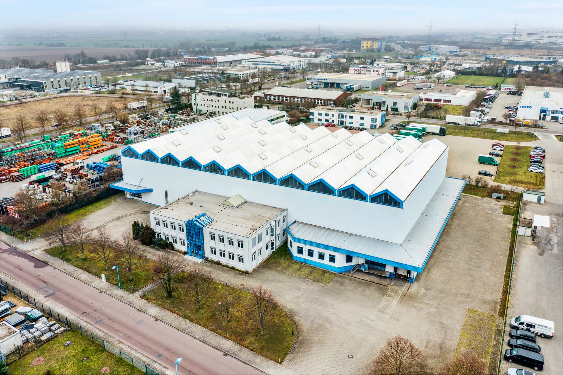 An aerial view of a large industrial warehouse with a white and blue facade, surrounded by smaller buildings, parking areas, and greenery in an industrial area.