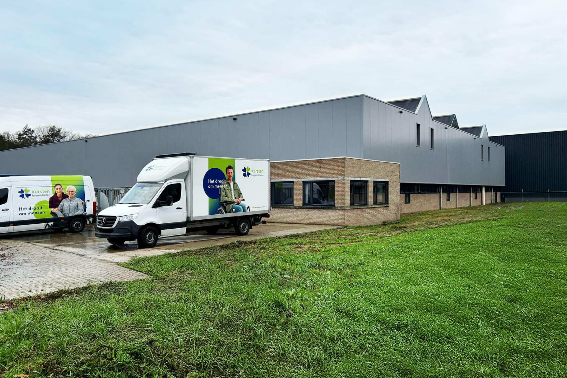 Two delivery trucks parked outside a large industrial building with gray siding on a cloudy day.