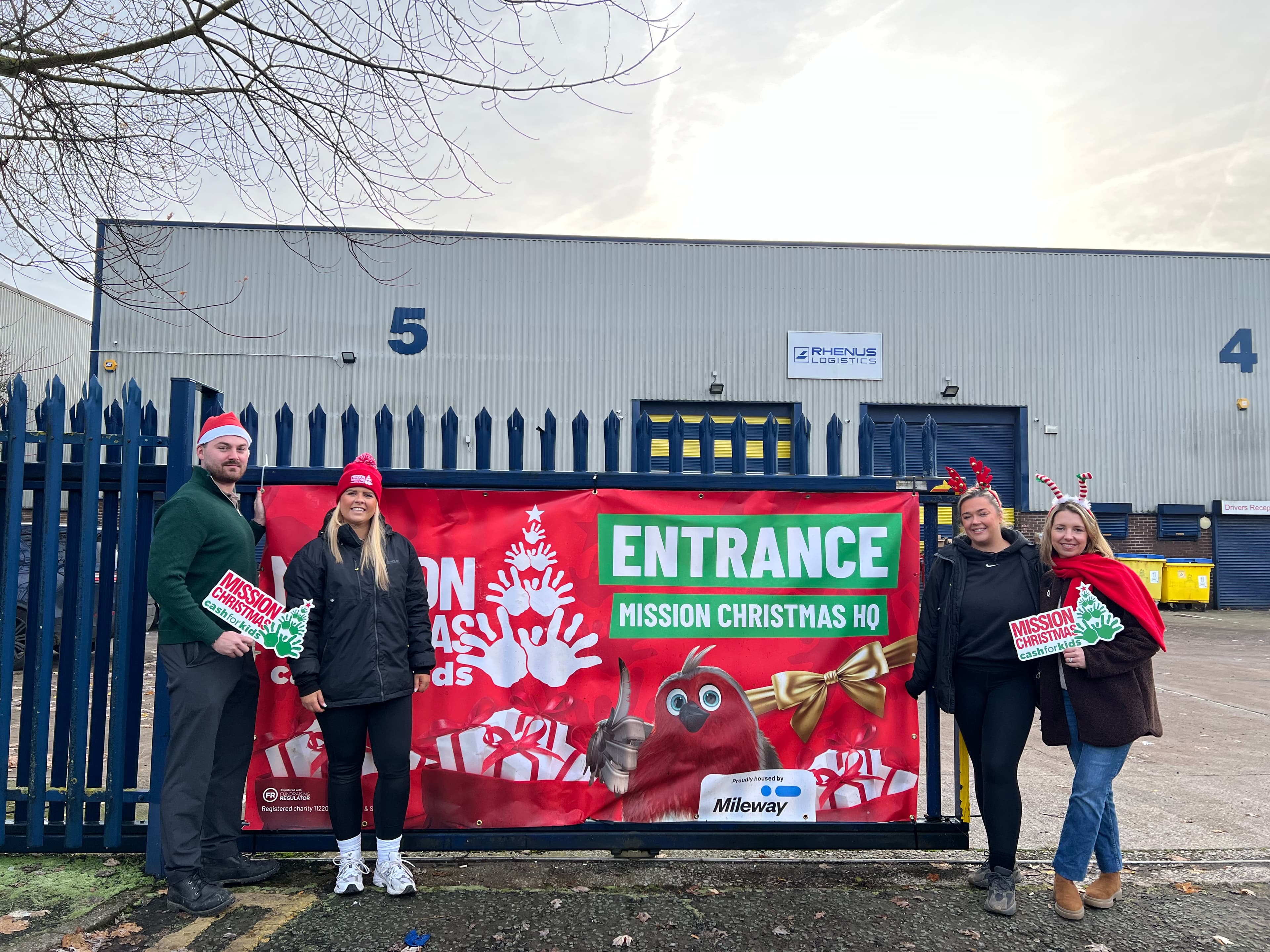 Four people stand in front of a building decorated for a Christmas mission event, holding festive signs, with one person wearing a Santa hat and another with reindeer antlers.