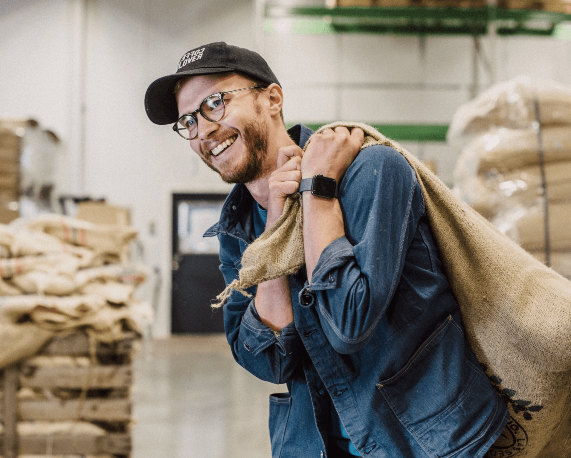 A man wearing glasses and a cap smiles while carrying a large burlap sack on his shoulder inside a warehouse with stacked bags in the background.