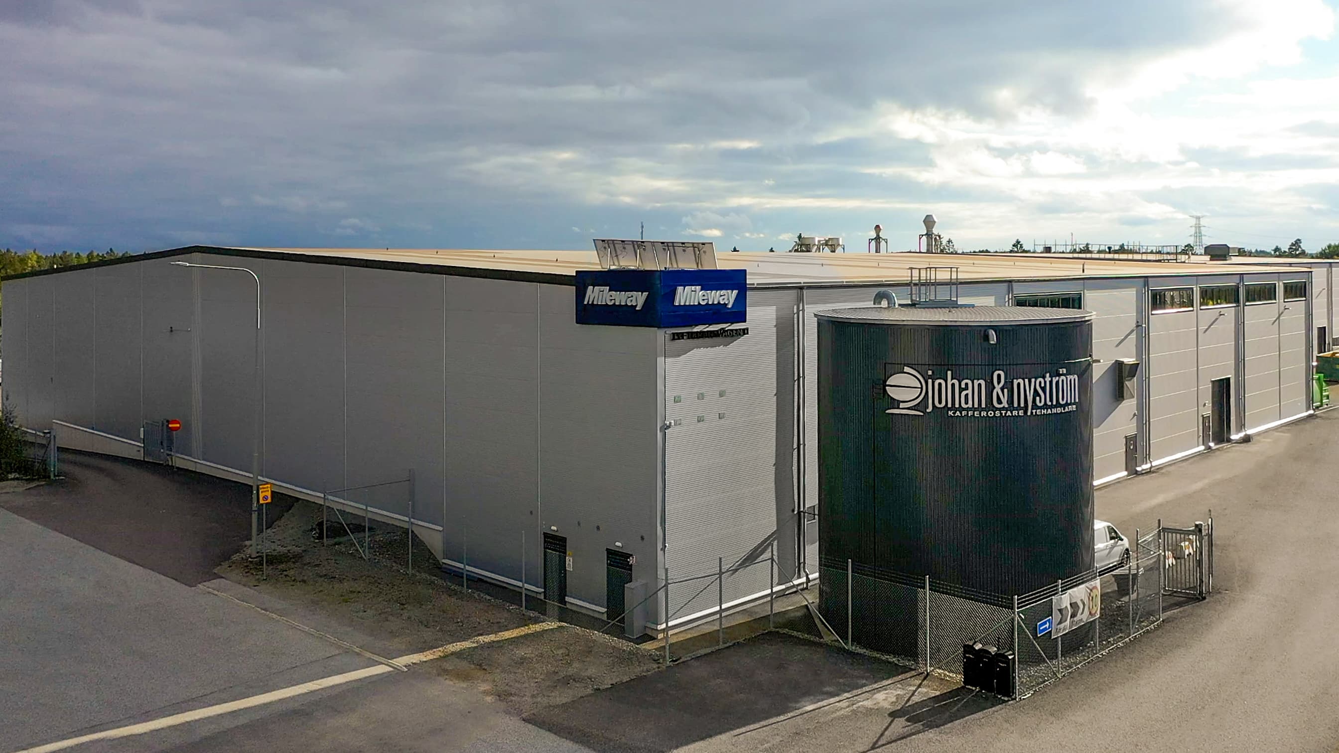 Large industrial building with Miljeway and Johan & Nyström signage, surrounded by a fenced area and adjacent road under a cloudy sky.