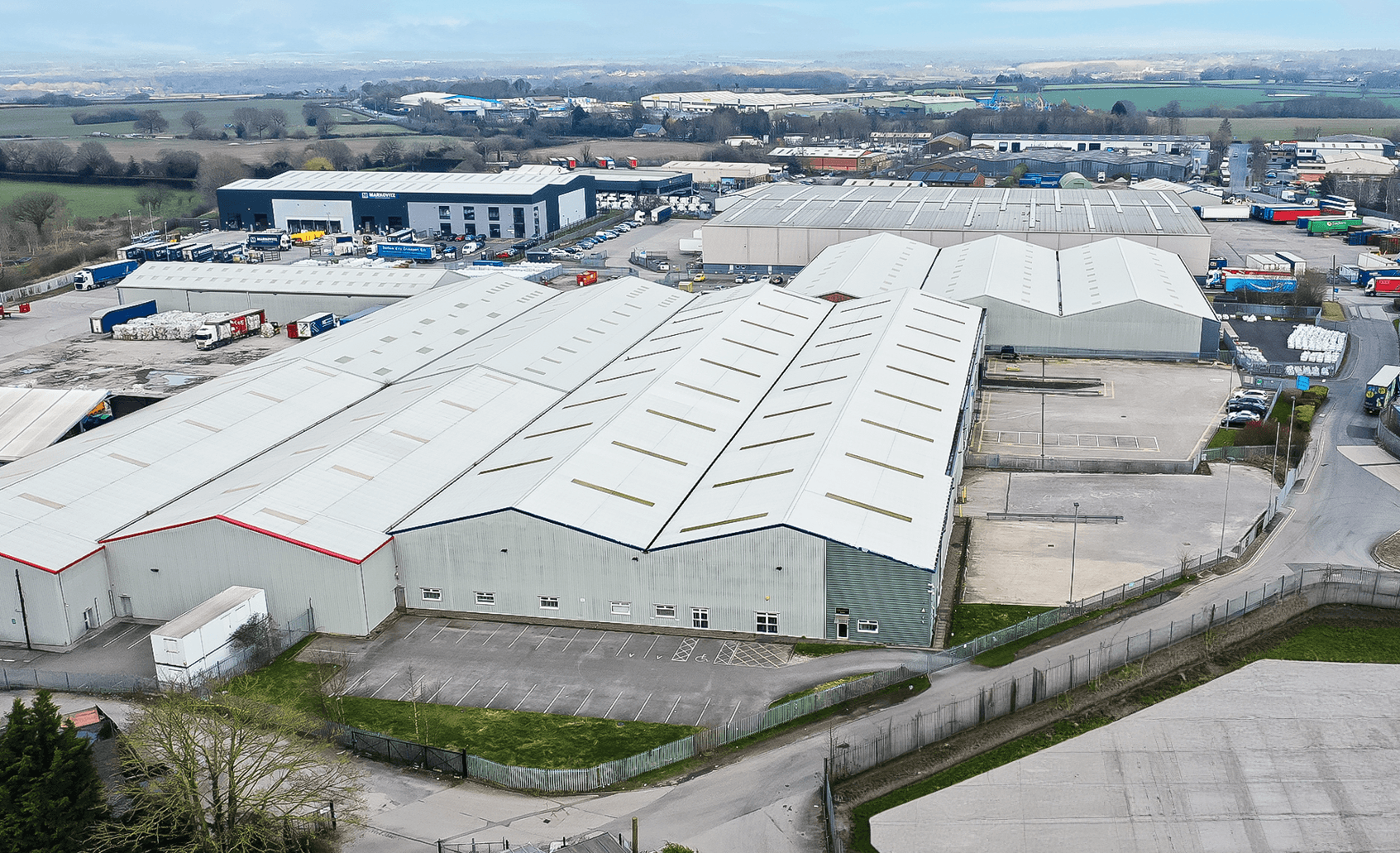 Aerial view of an industrial complex with multiple large warehouse buildings, parking areas, and surrounding green fields under a cloudy sky.