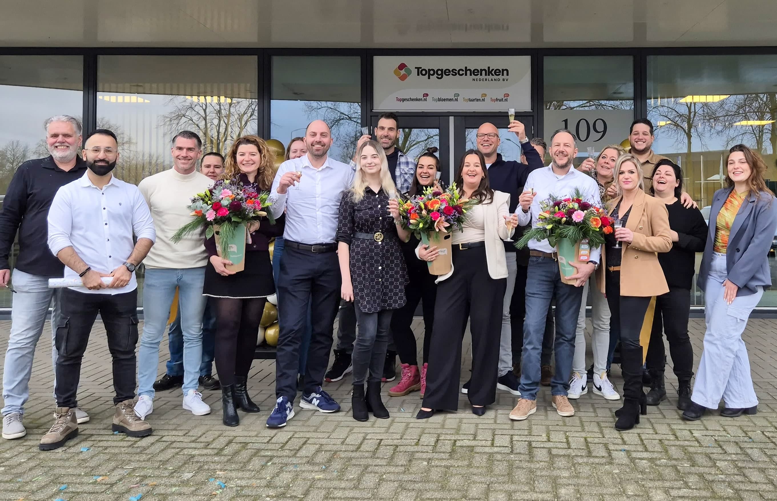 A group of people standing outside a building, some holding bouquets of flowers, smiling for a group photo. The building has a sign reading Topgeschenken.