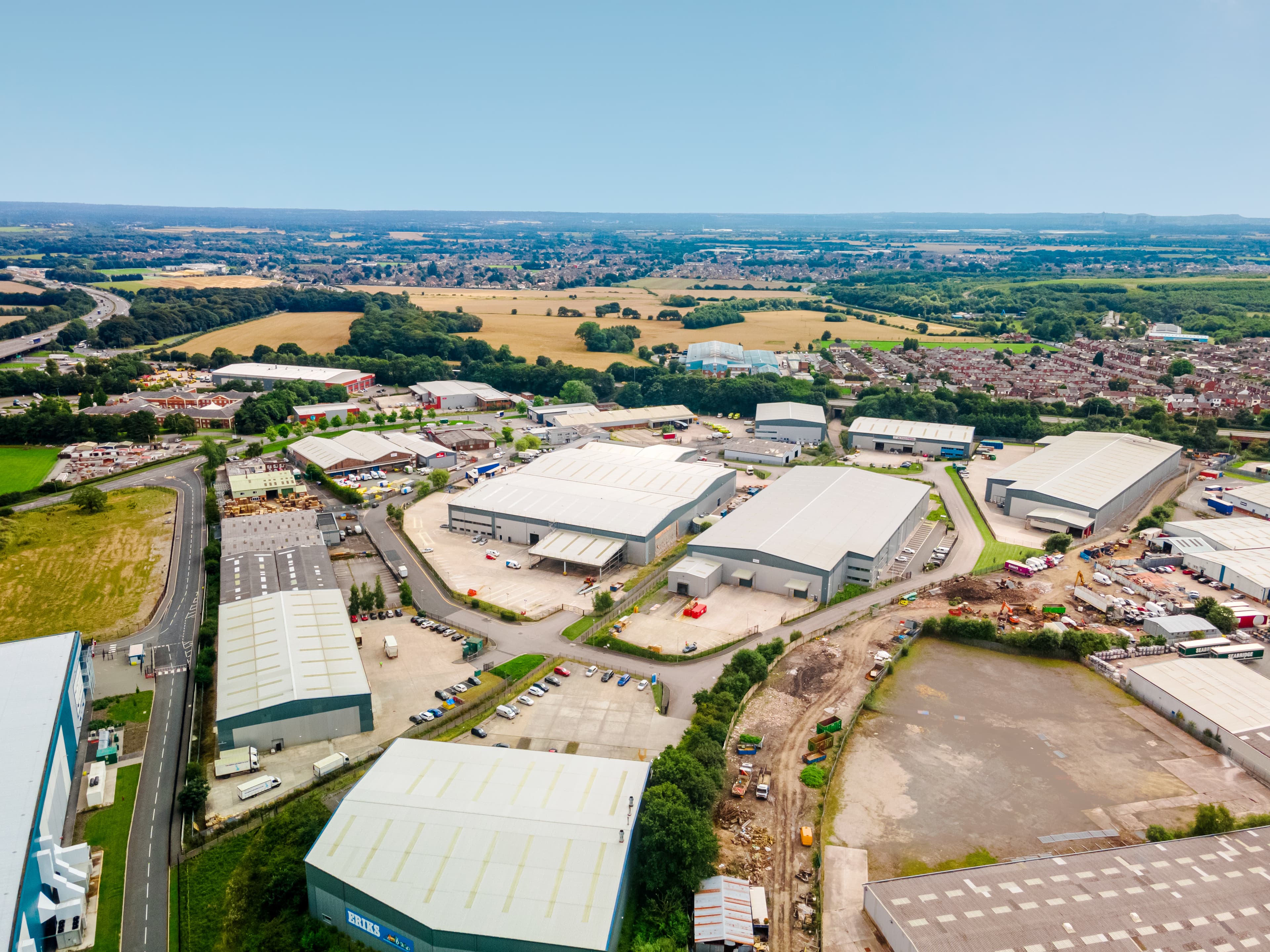 Aerial view of an industrial complex with warehouses, surrounding roads, and nearby residential and agricultural areas under a clear sky.