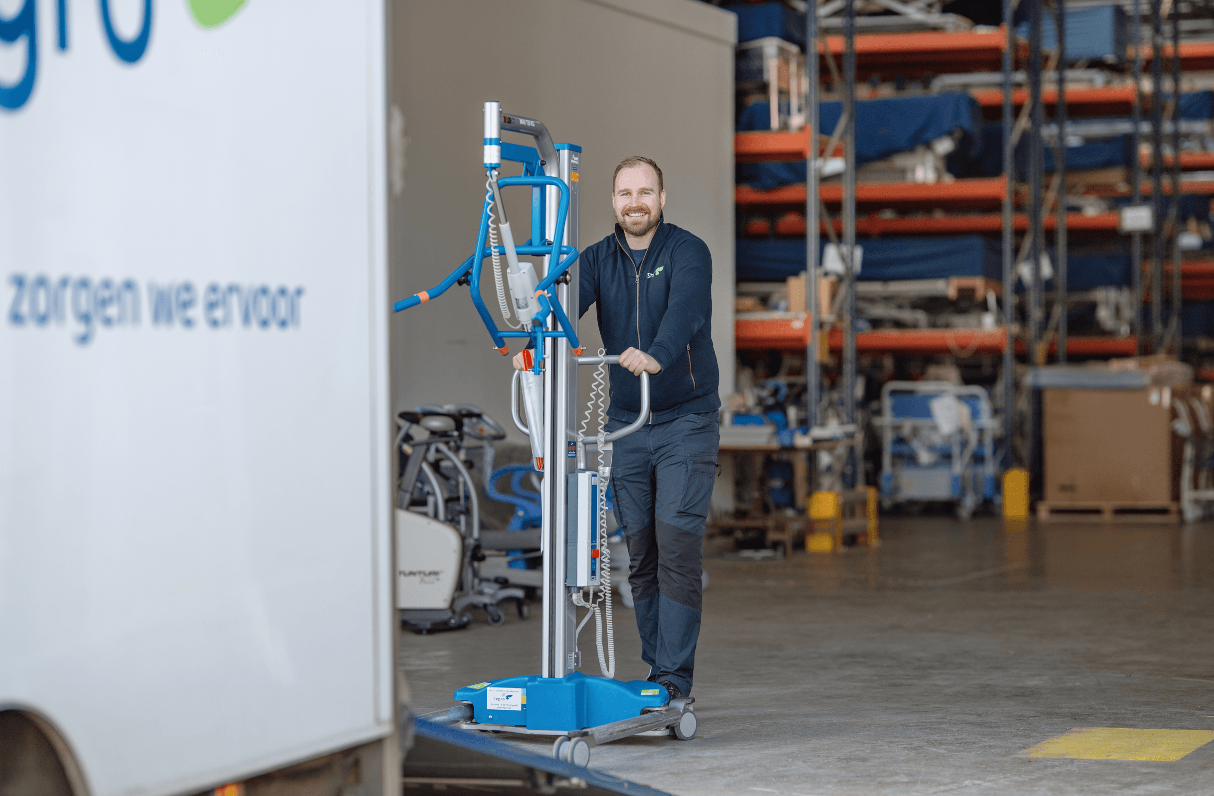 A person in a warehouse moves a large piece of equipment on wheels near a truck, with shelves and various items in the background.