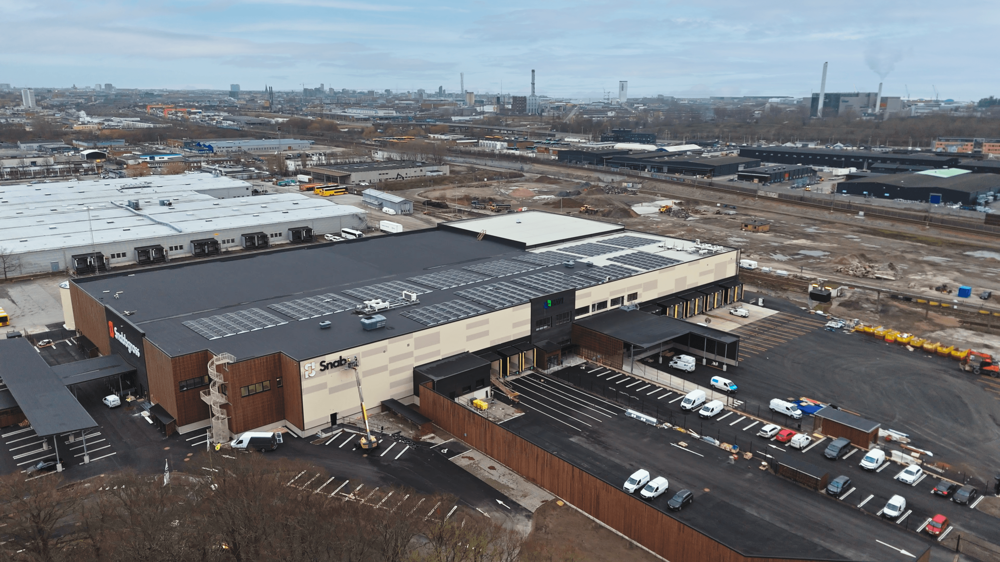 Aerial view of a large industrial building with a marked parking lot, surrounded by an urban landscape and construction areas under a cloudy sky.