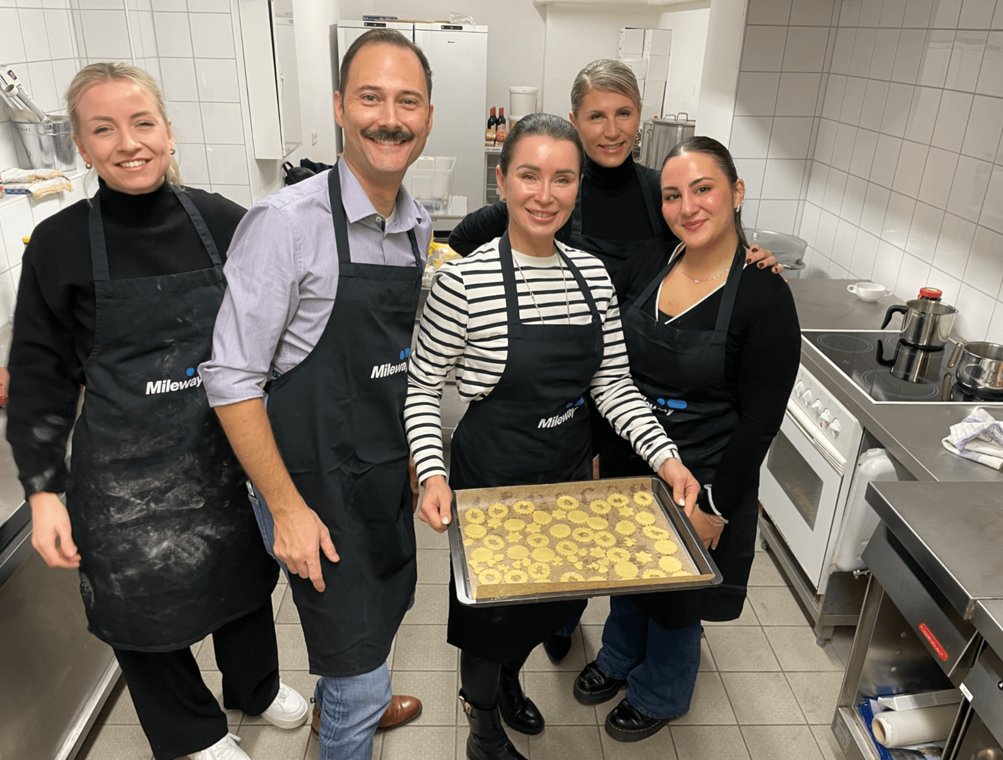 Five people in aprons stand in a kitchen, smiling, with one holding a tray of cookies ready for baking.