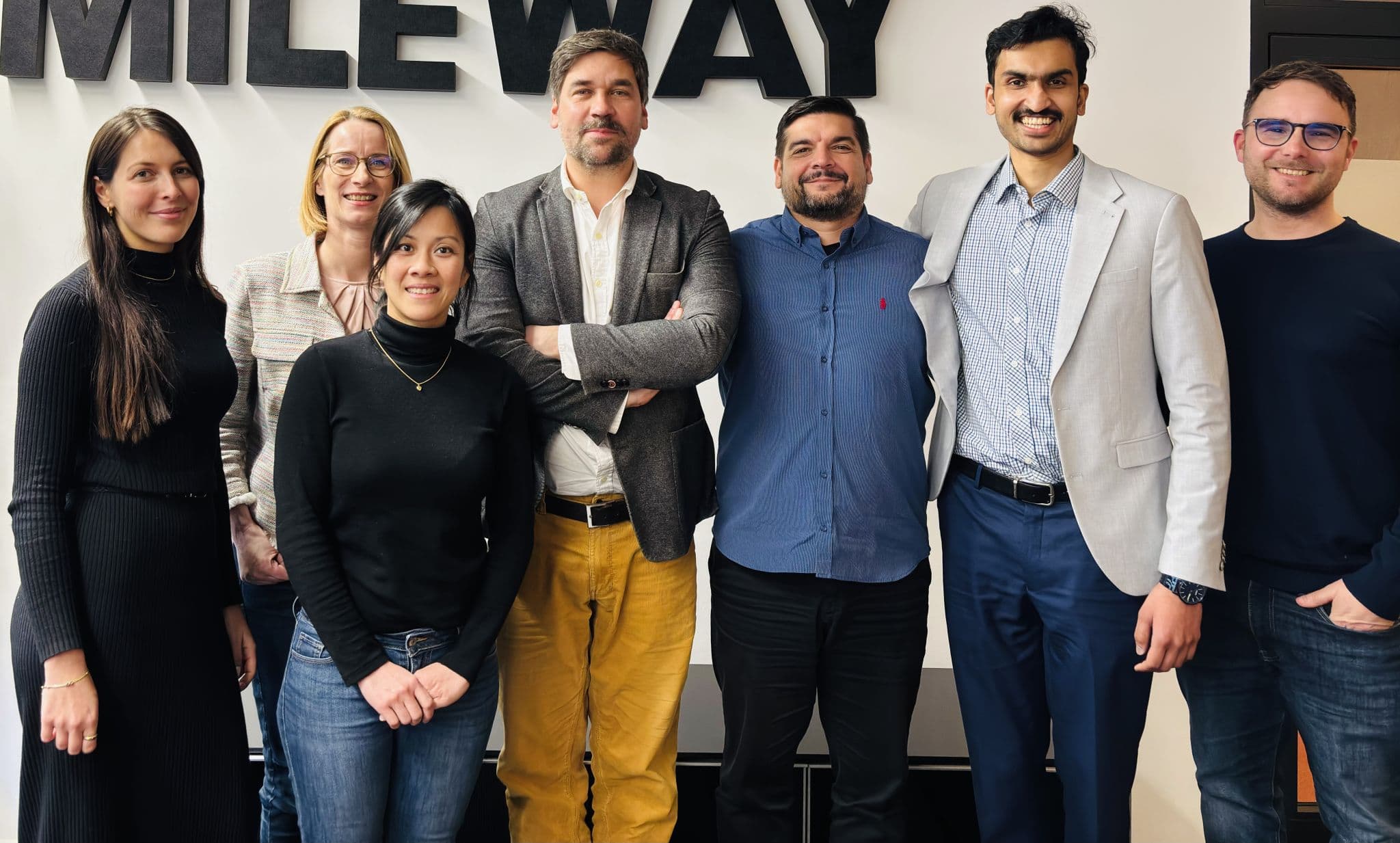 Seven people standing side by side in front of a wall with the partial word MILEWAY visible above them.
