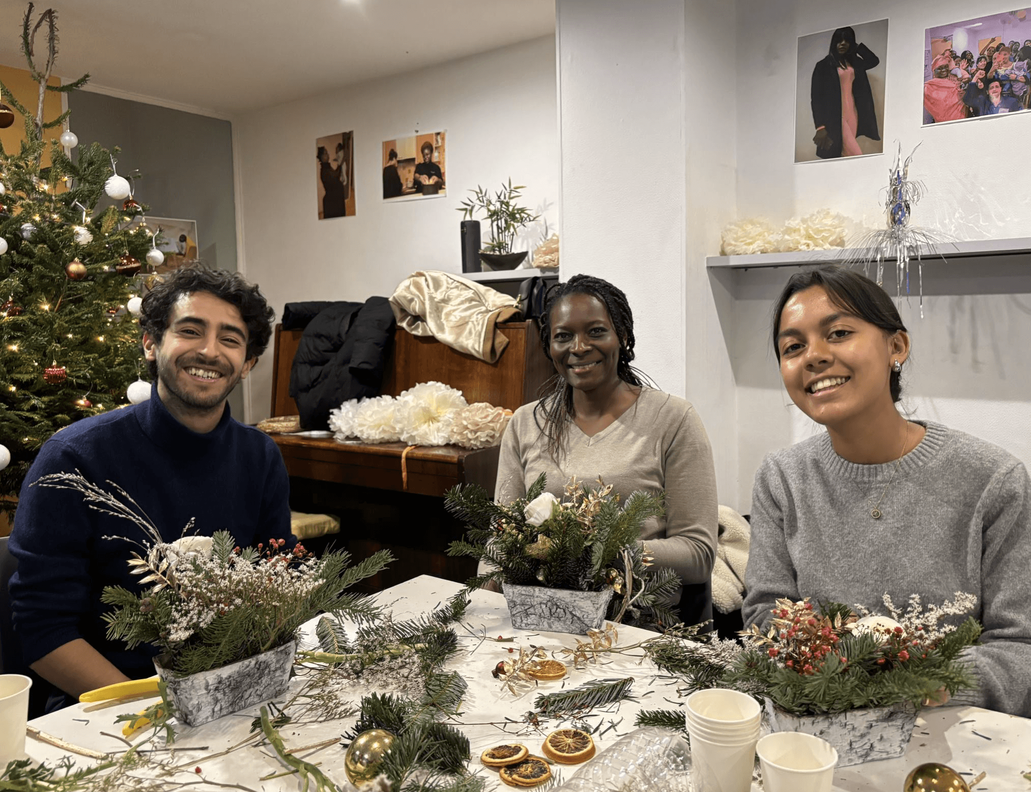 Three people smile while sitting at a table with holiday decorations, including arrangements of greenery and berries, in a room with a Christmas tree and photos on the wall.