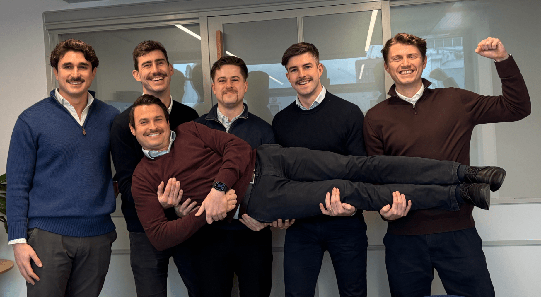 Six men in a room, five standing and one being held horizontally by the others. They are all smiling and have mustaches.