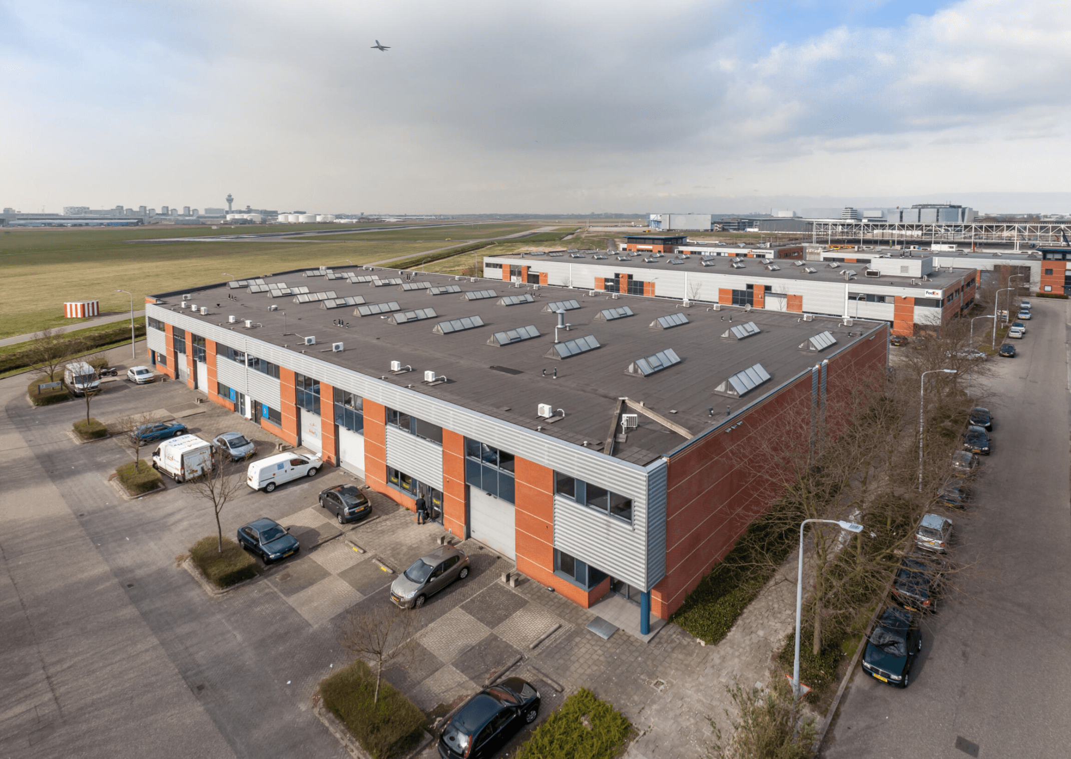 Aerial view of industrial buildings with parking lots and vehicles. An airplane is visible in the sky above green fields.