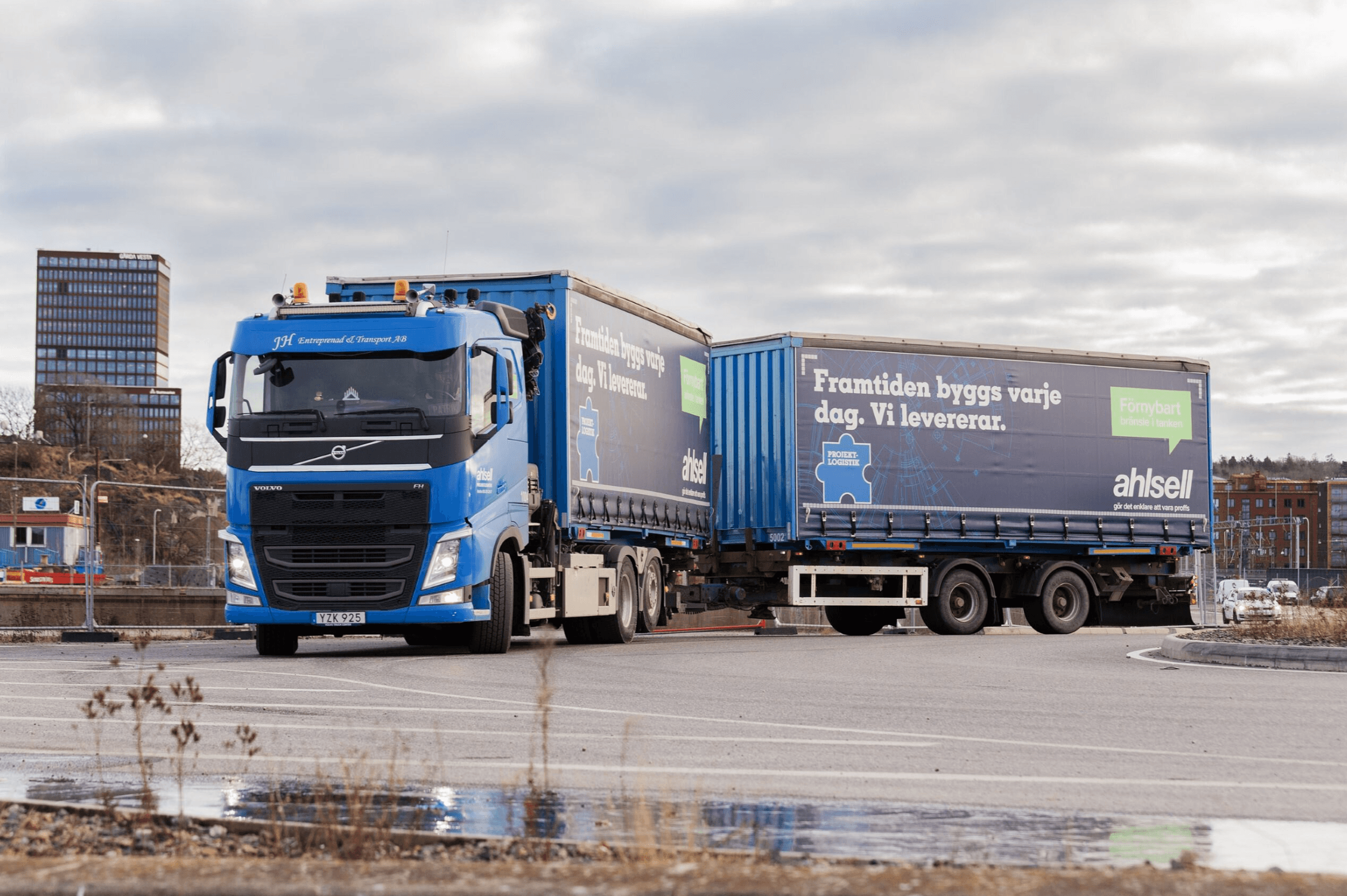 Blue Volvo semi-truck with attached trailer parked in an industrial area. The trailer features company branding and text in Swedish. Cloudy sky and buildings in the background.