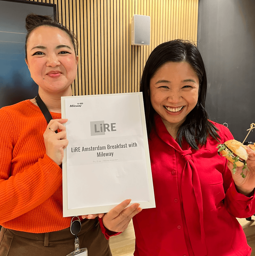 Two people smiling; one holds a LiRE Amsterdam Breakfast with Mileway sign, the other holds a sandwich.