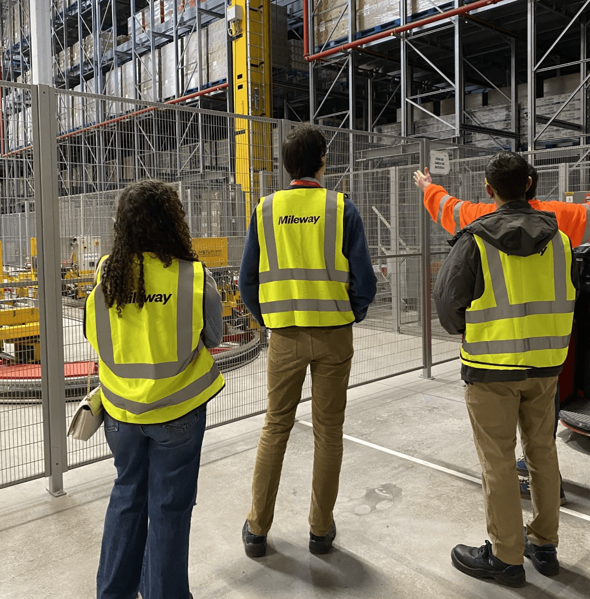 Three people in high-visibility vests stand in a warehouse, facing a person who is gesturing towards storage shelves.