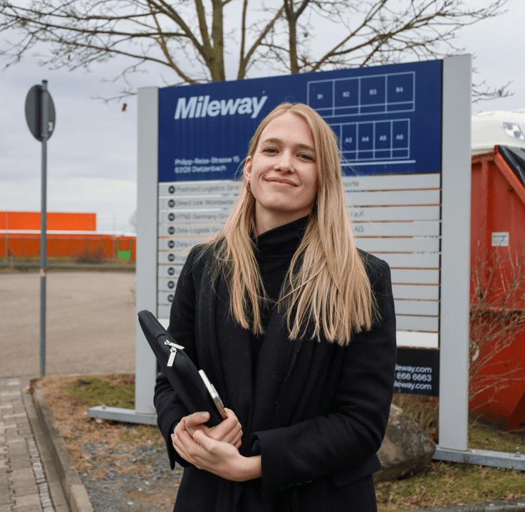 Woman in black coat holding a folder stands in front of a Mileway site sign outdoors.