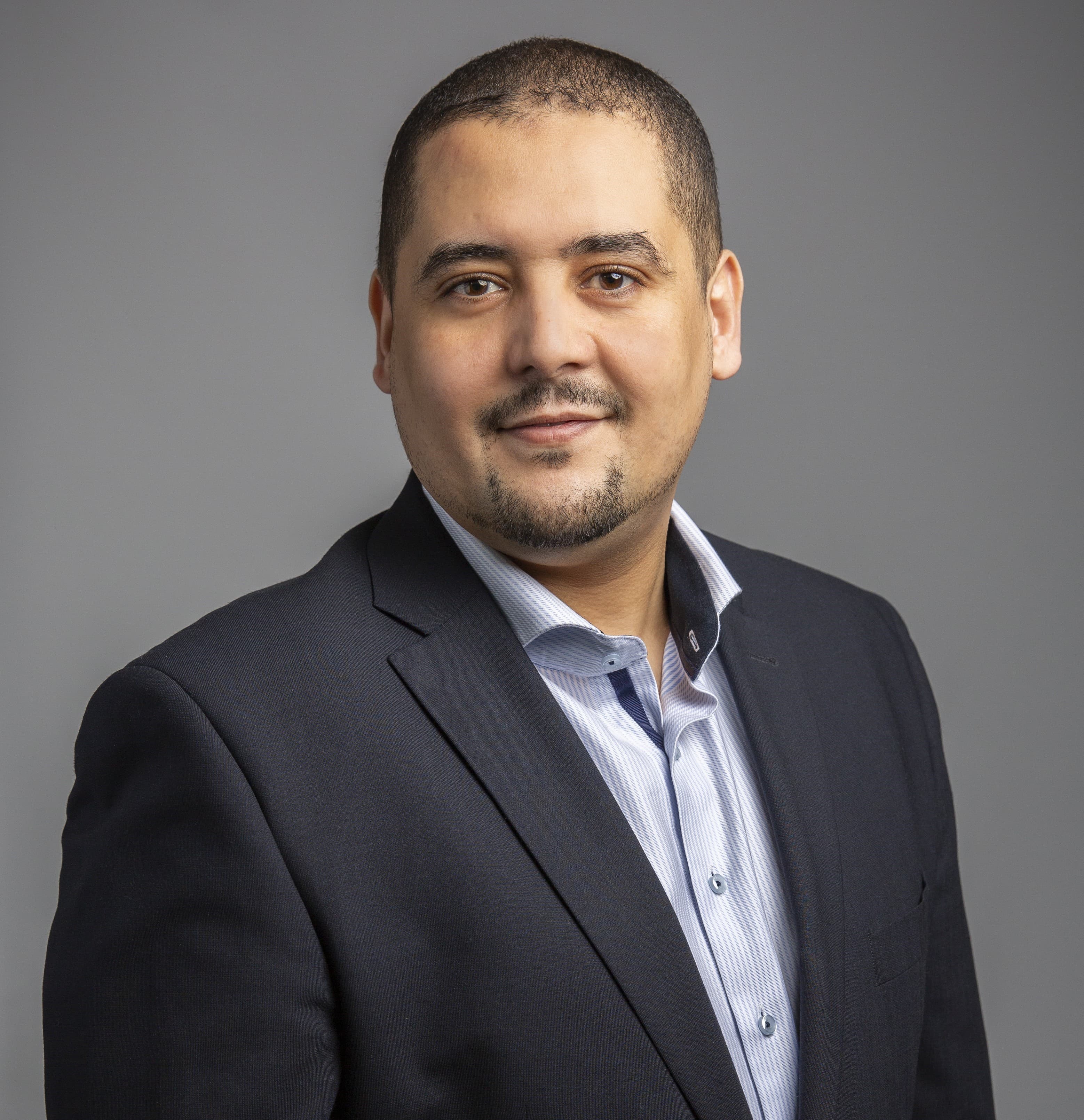 A person in a dark suit jacket and light striped shirt poses for a professional portrait against a plain gray background.