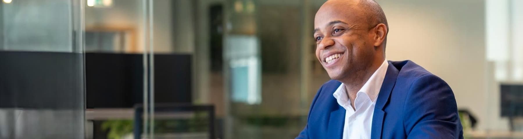 Man in a blue suit smiling in a modern office setting with glass walls and greenery.