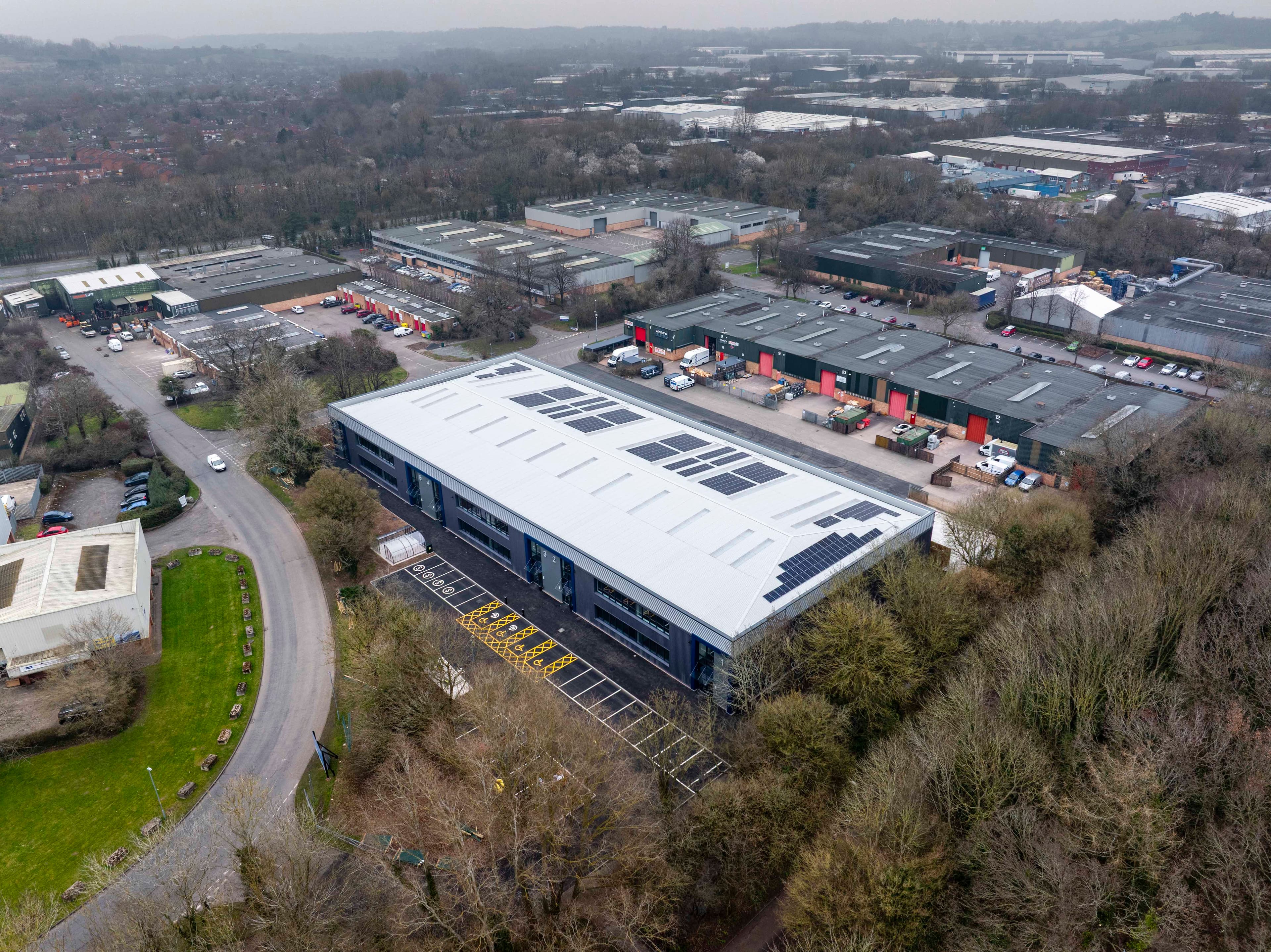 Aerial view of a large industrial building with solar panels on the roof, surrounded by other buildings, trees, and roads on an overcast day.
