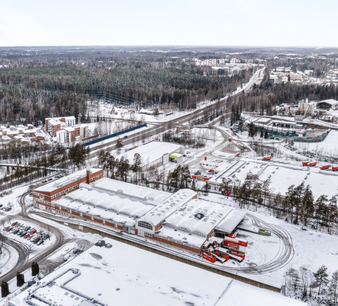 Aerial view of a snowy industrial area with several rectangular buildings and red trucks, surrounded by trees and distant residential areas.