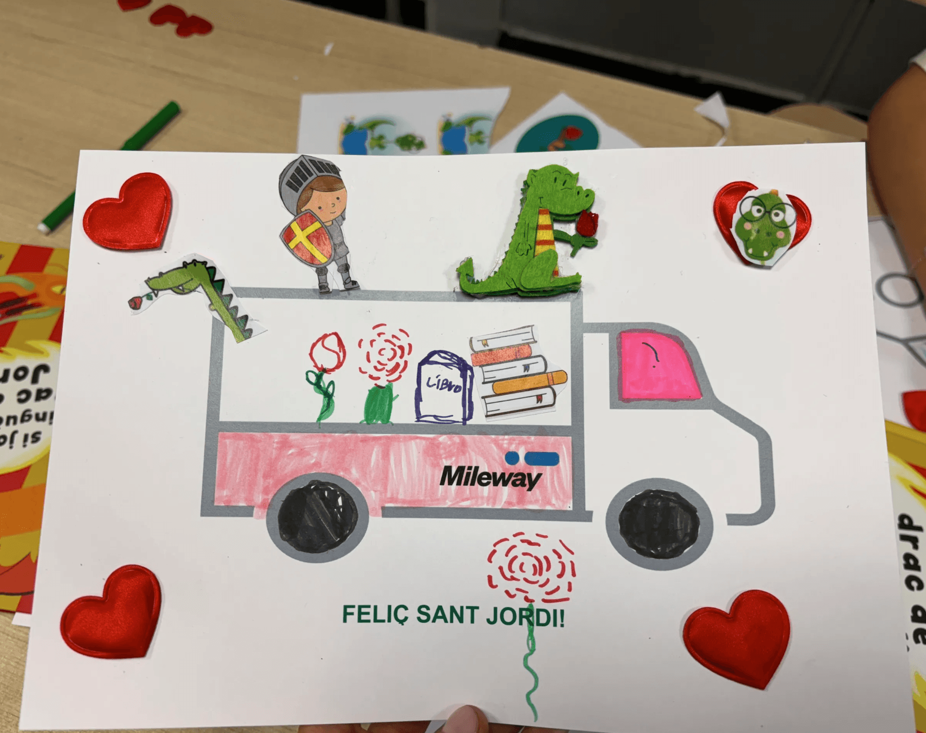 A handmade card shows a truck with a knight, dragon, books, roses, and heart stickers. Text reads Mileway on the truck and FELIC SANT JORDI! at the bottom.