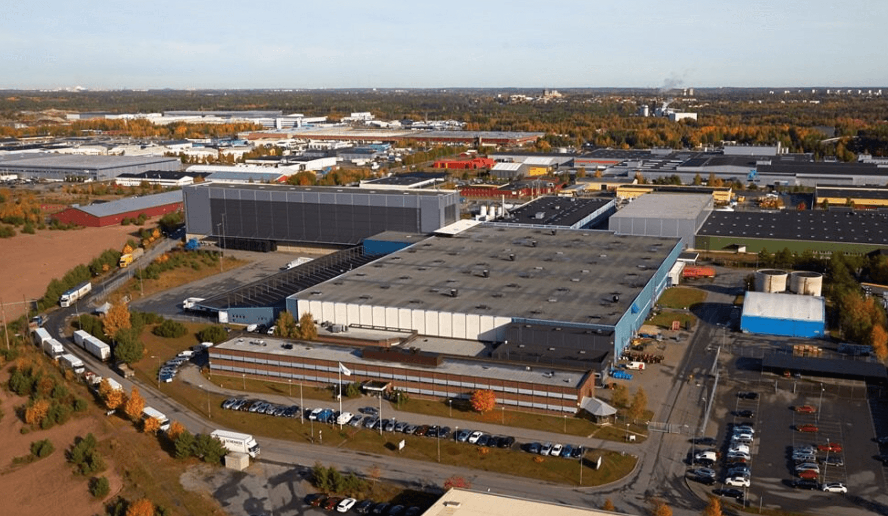 Aerial view of an industrial complex with large warehouse buildings, parking areas, and surrounding roads, set amidst a semi-urban landscape.