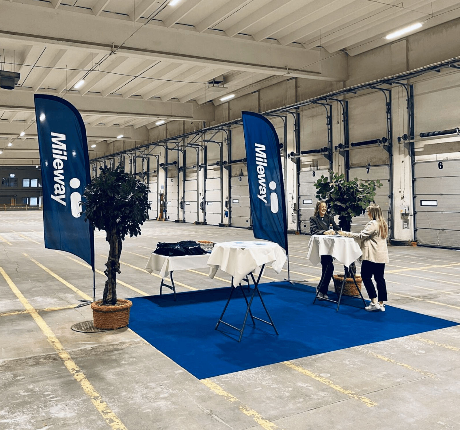 Two people converse at a table on a blue mat in a large, empty warehouse with Mileway banners and potted plants nearby.