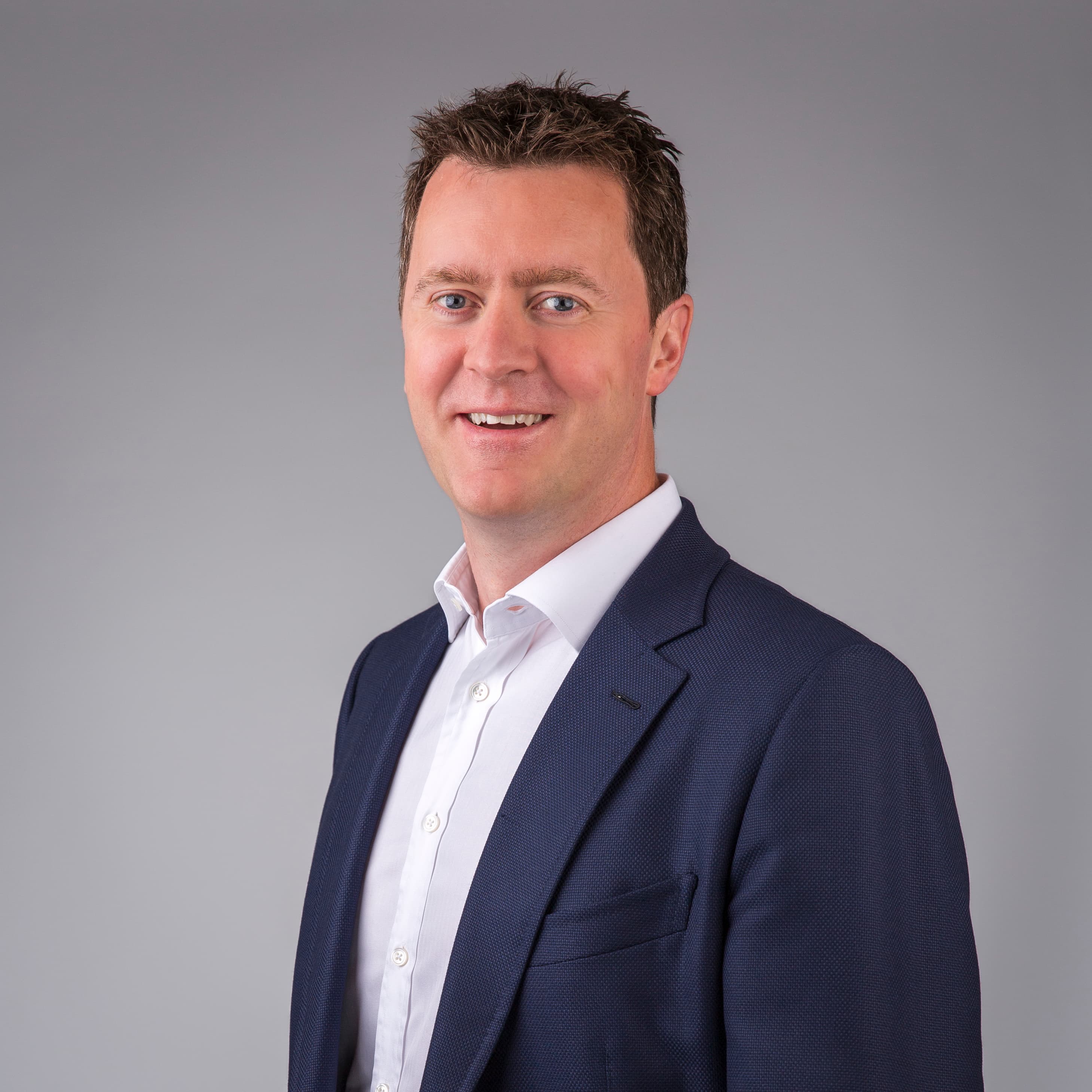 A person in a dark suit jacket and white shirt is smiling, standing in front of a plain gray background.