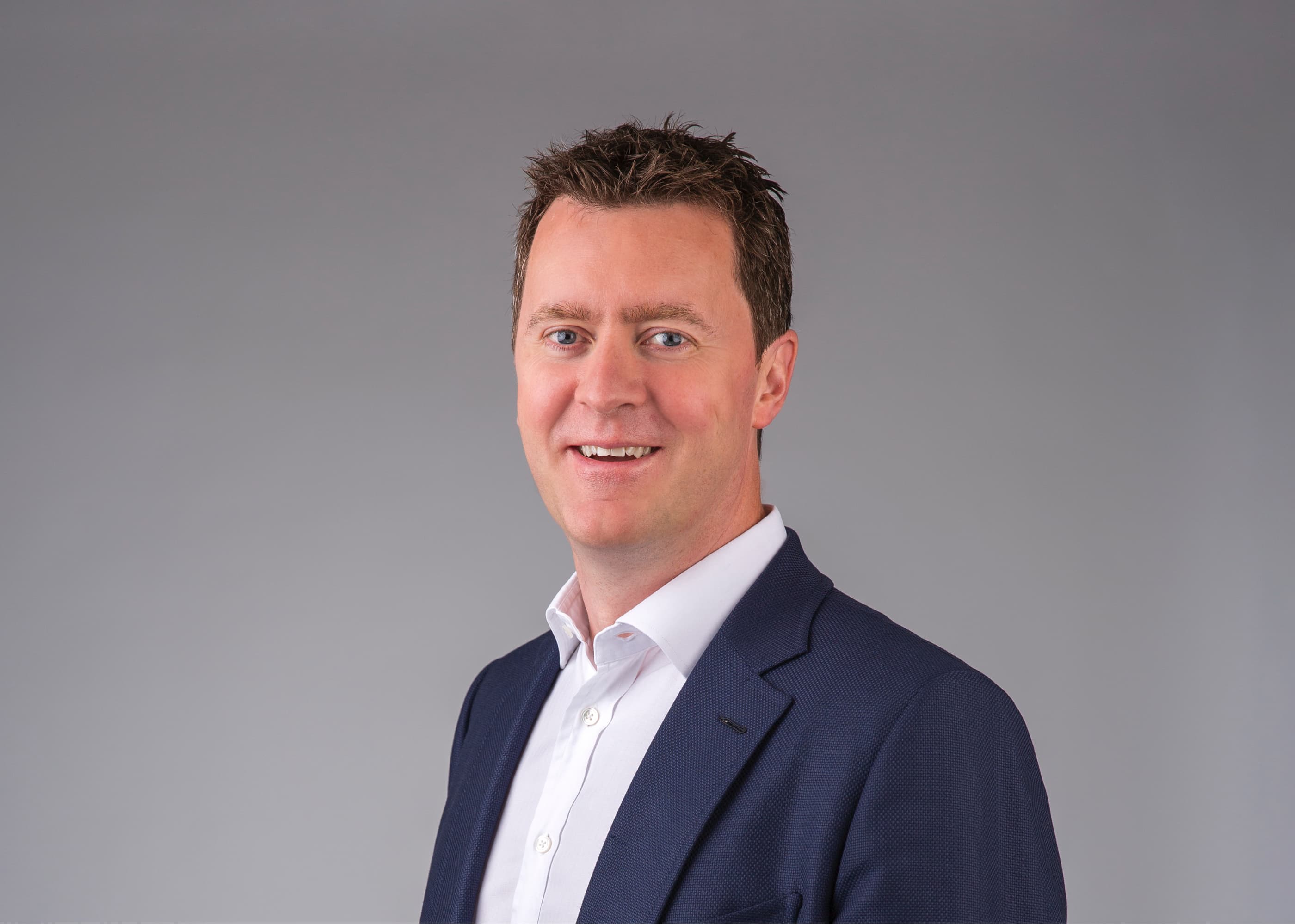 A man in a navy blue suit and white shirt smiles at the camera against a plain gray background.
