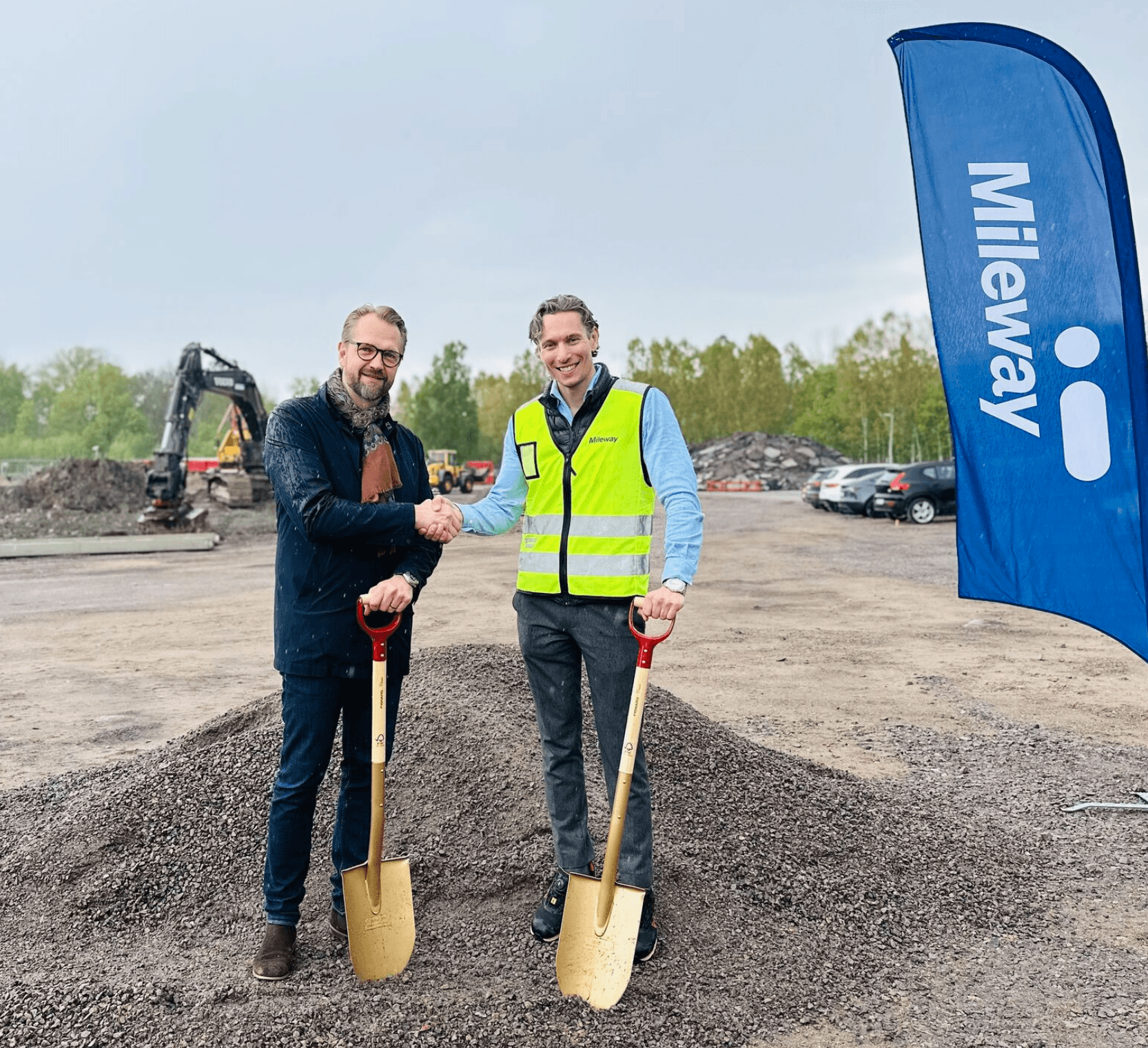 Two men stand on gravel, shaking hands and holding gold shovels at a groundbreaking site near a Mileway flag, with construction equipment and cars in the background.