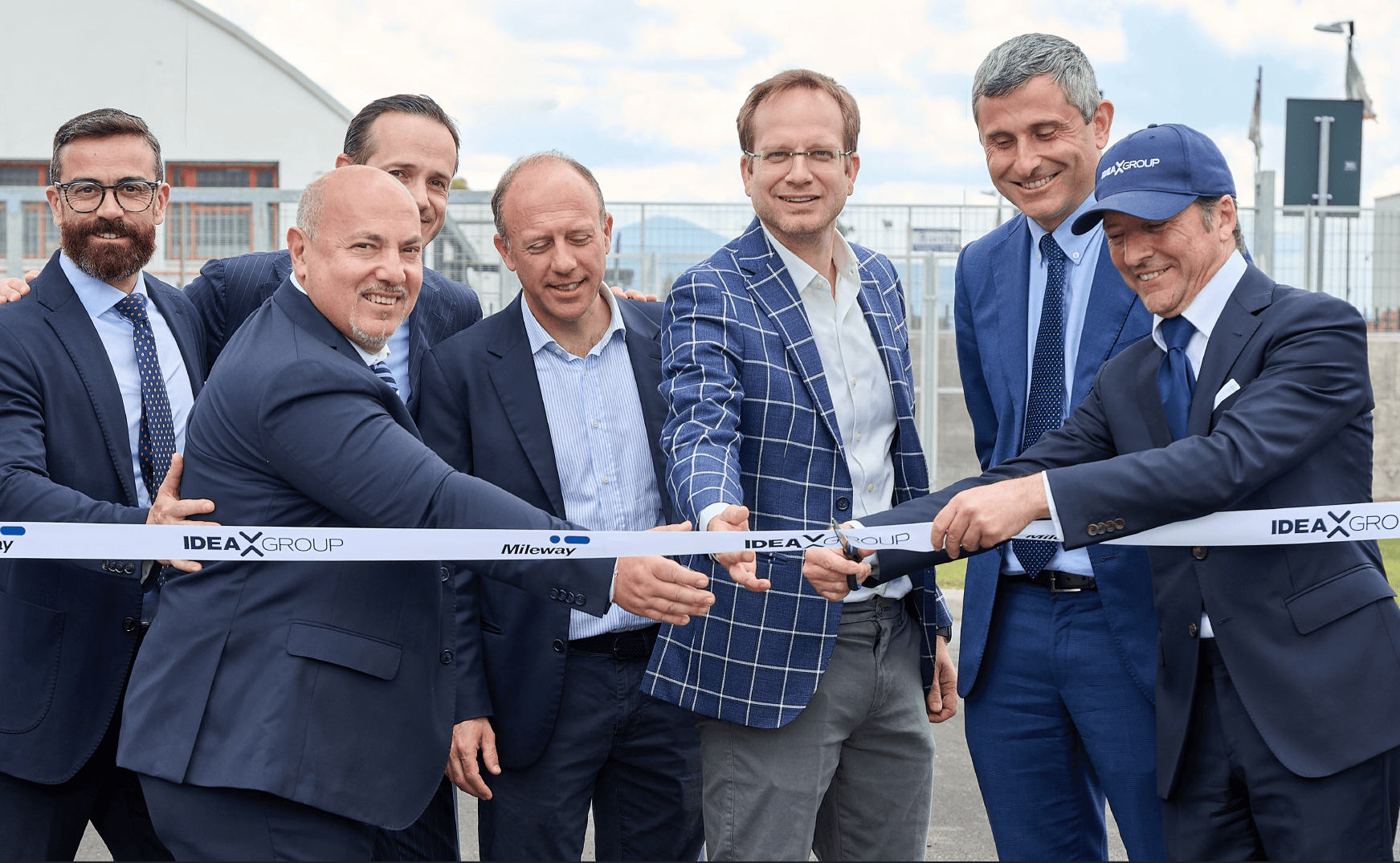 Seven men in business attire smile and cut a ribbon together at an outdoor event, signifying the opening or launch of a new facility or project.