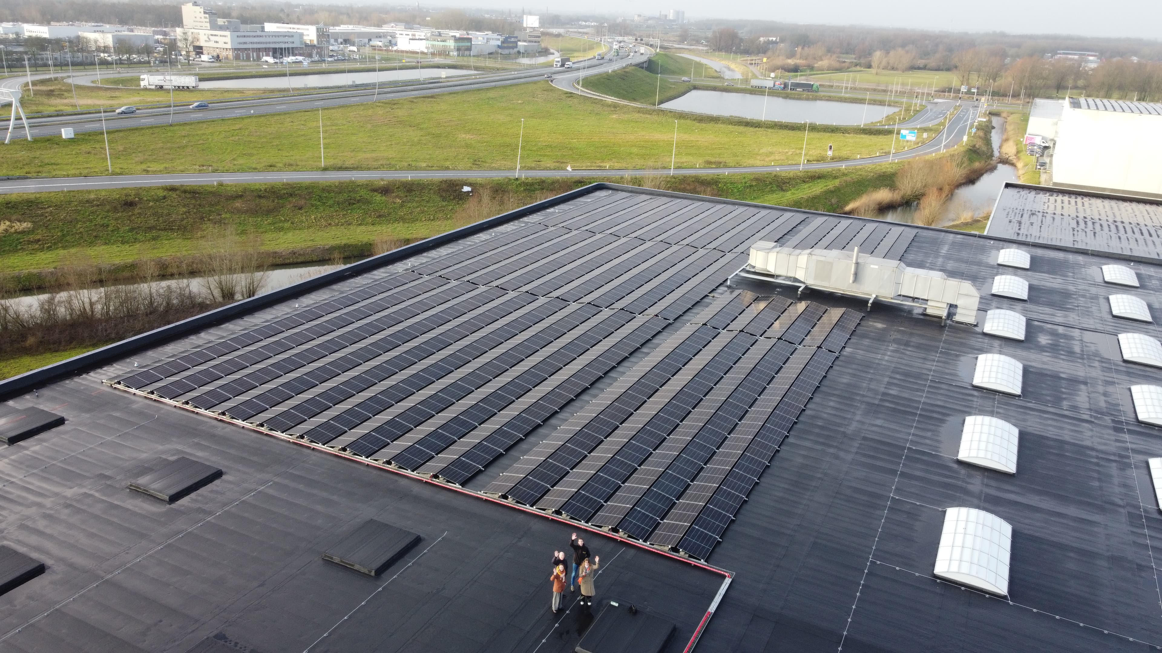 A large array of solar panels installed on the flat roof of an industrial building, with two people standing near the edge and roads visible in the background.