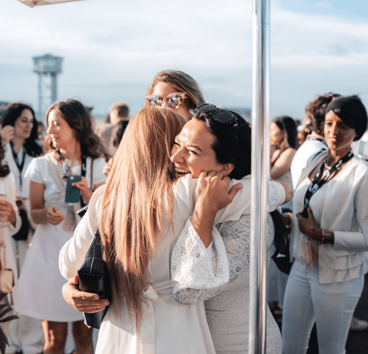 Two women embrace and smile at an outdoor event; several other people dressed in white are mingling in the background.