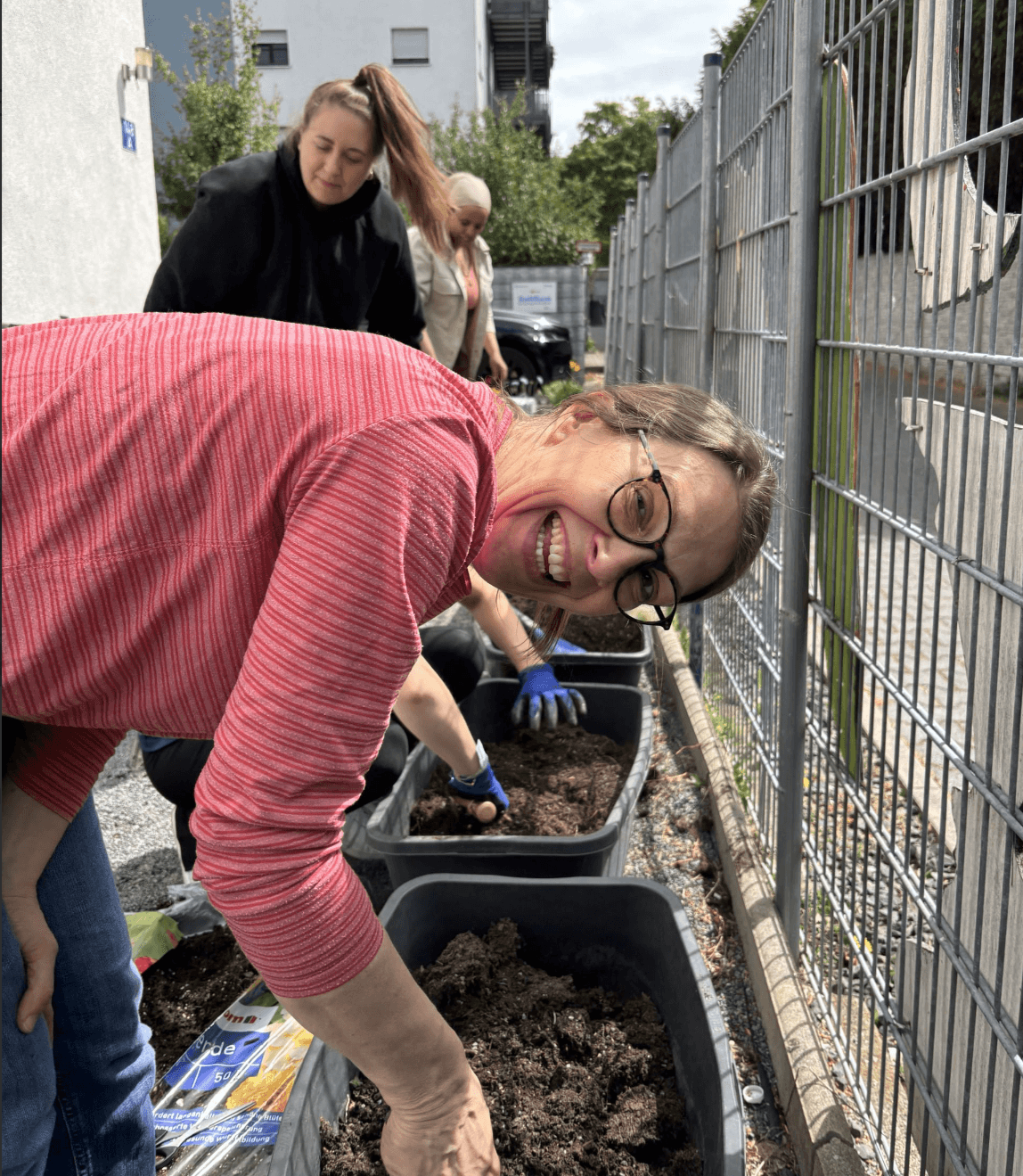 A woman in glasses smiles at the camera while gardening in soil containers alongside two other people near a metal fence.