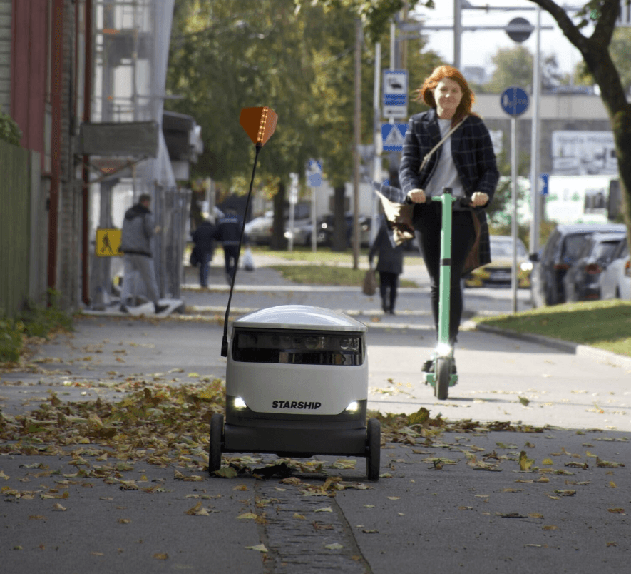 A delivery robot labeled Starship moves along a sidewalk while a woman on an electric scooter approaches from behind.