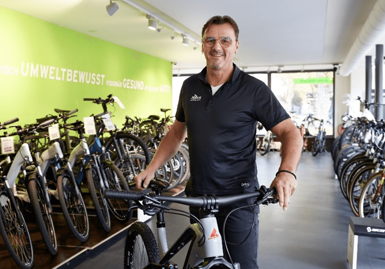 A man stands in a bicycle shop, holding a bike, with other bicycles displayed in the background and a green wall with German text.