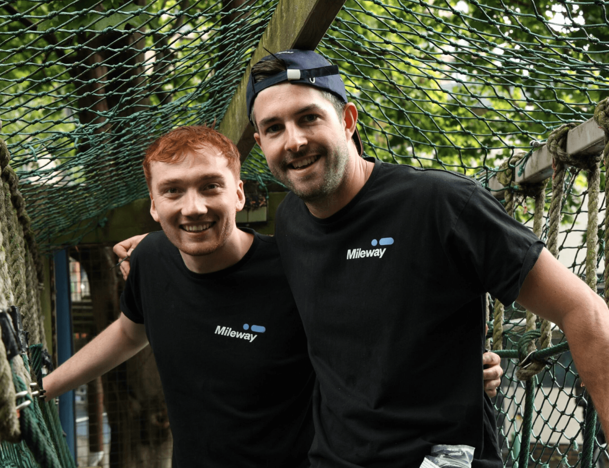Two men wearing black Mileway shirts pose and smile on a rope bridge outdoors, surrounded by trees and netting.
