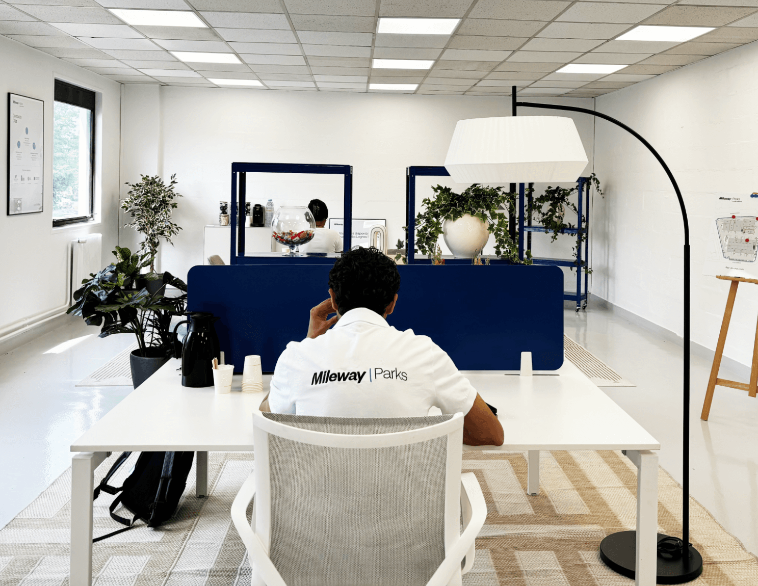 A person sits at a white desk in a modern office, facing away. The desk has cups and a phone. Blue dividers, plants, and office decor are visible in the background.