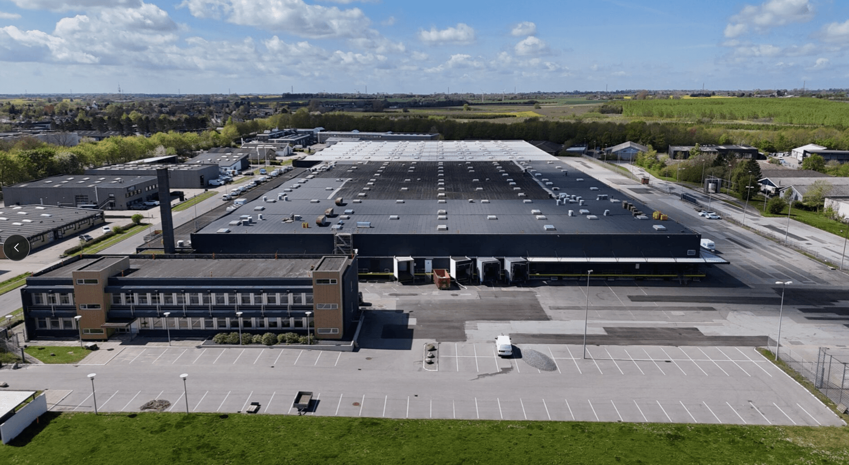 Aerial view of a large industrial warehouse with an attached office building, surrounded by parking lots and greenery under a partly cloudy sky.