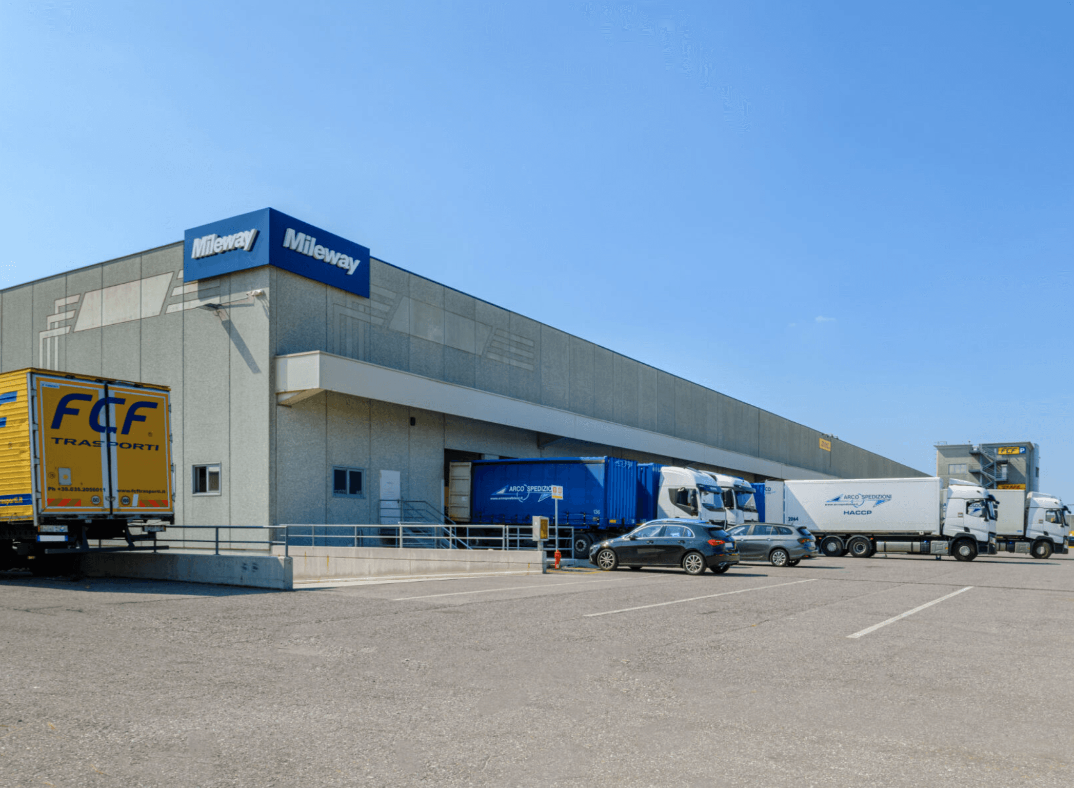 A logistics warehouse with Mileway signage, several delivery trucks, and a few parked cars in front under a clear blue sky.