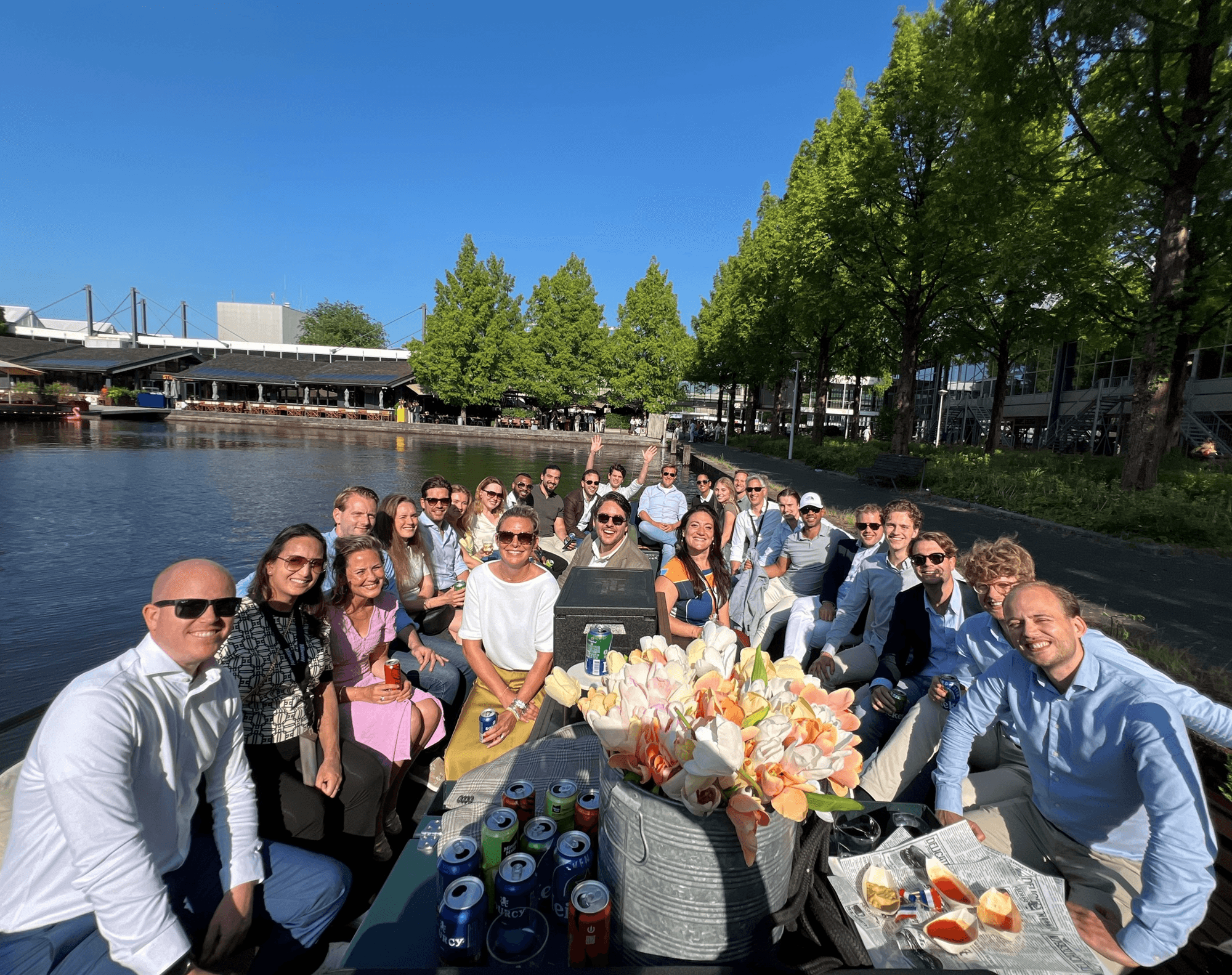 A large group of people are sitting on a boat docked by a tree-lined canal, smiling at the camera on a sunny day. Drinks and snacks are on the table in front of them.