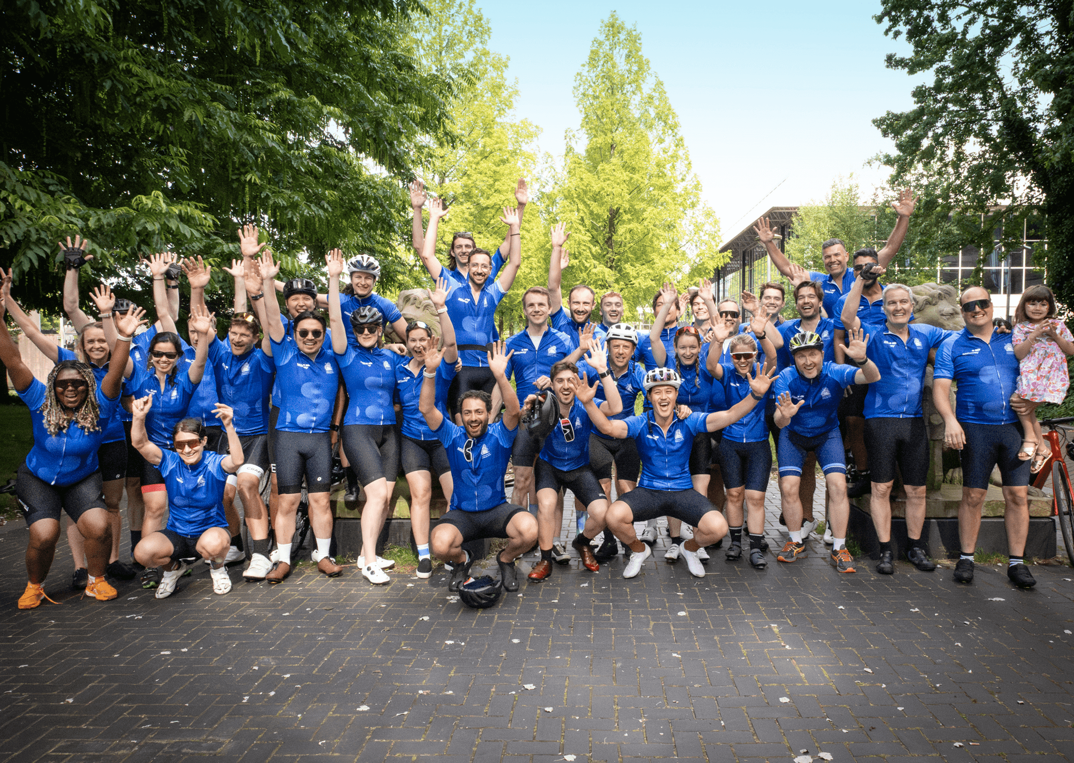 A large group of cyclists in matching blue jerseys pose outdoors with raised arms, smiling for a group photo on a paved path surrounded by greenery.