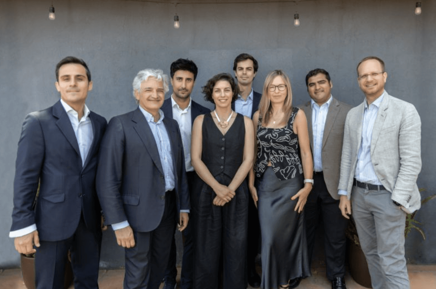 Eight professionally dressed people stand together outdoors in front of a gray wall, posing for a group photo under string lights.