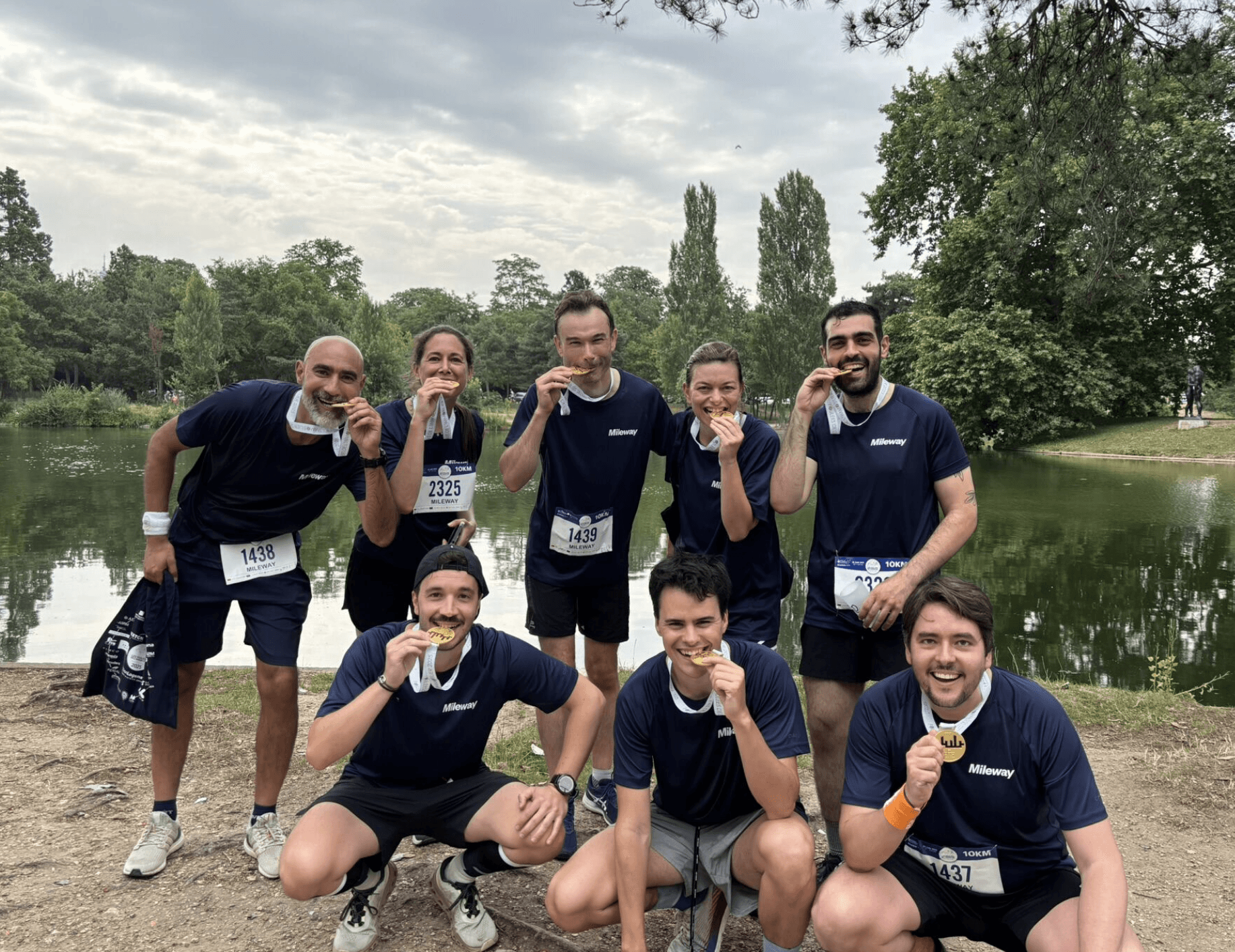 Eight people wearing navy blue shirts pose by a lake, holding medals and smiling, appearing to celebrate after a race or marathon.
