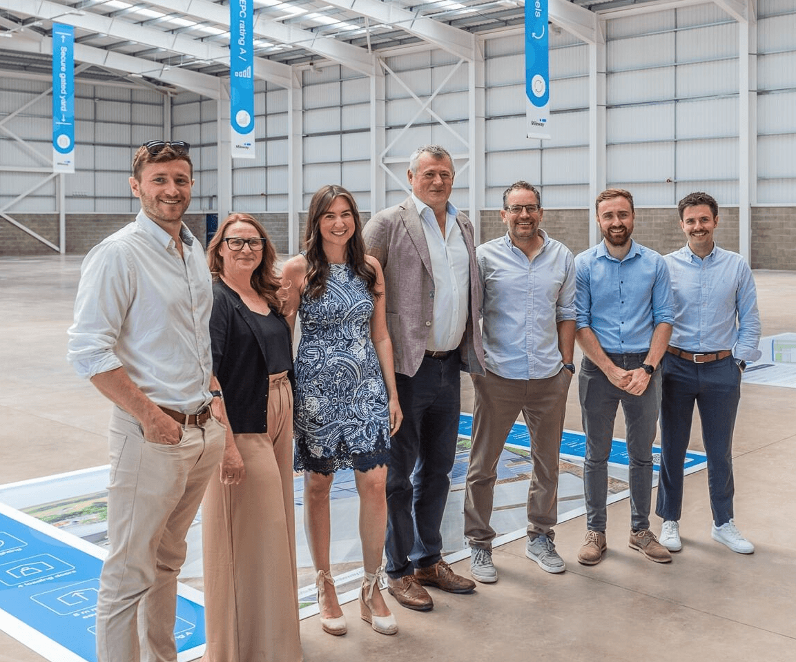 Seven people standing in a row inside a large, bright industrial warehouse, with informational banners and floor displays visible in the background.