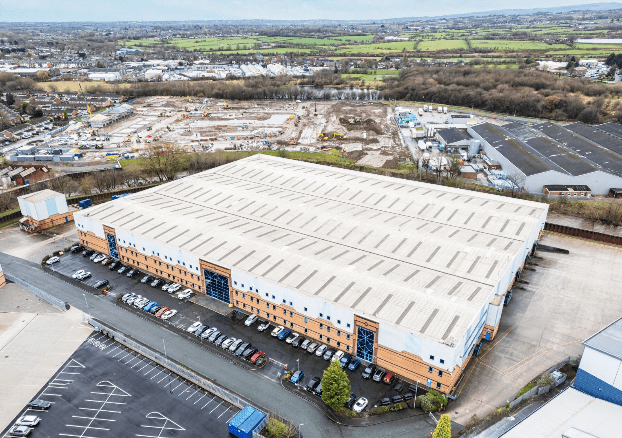 A large rectangular warehouse with a white roof is surrounded by parking lots and construction sites, with a town and fields visible in the background.