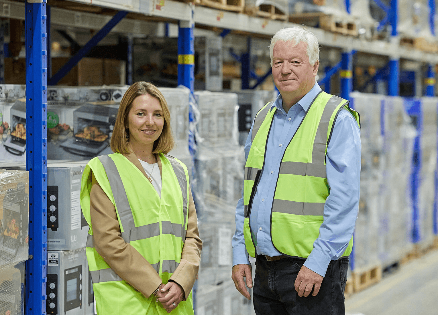 Two people wearing yellow safety vests stand in a warehouse aisle with shelves stocked with boxed products and pallets wrapped in plastic.